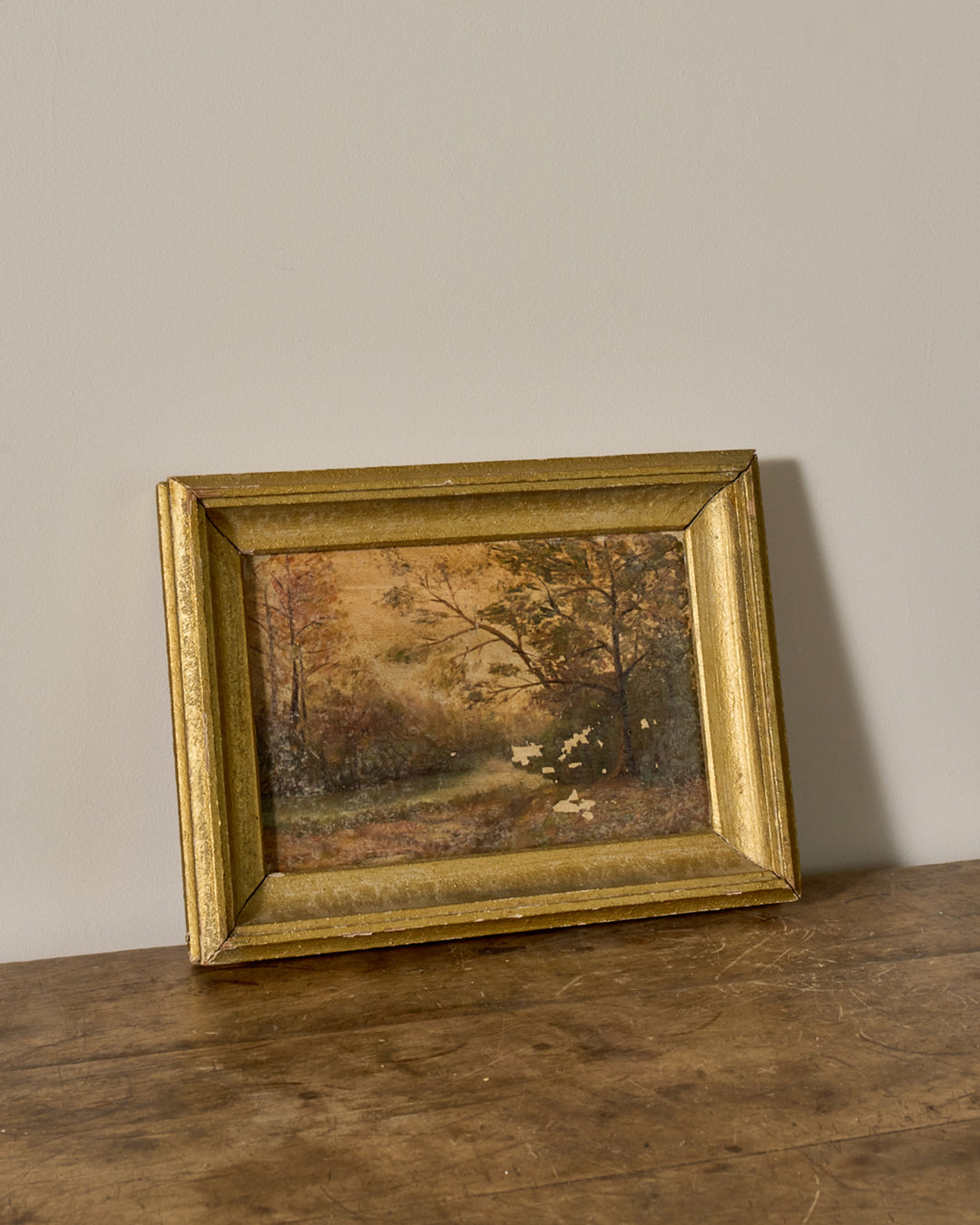 Gold-framed painting of a wooded landscape on a wooden table.