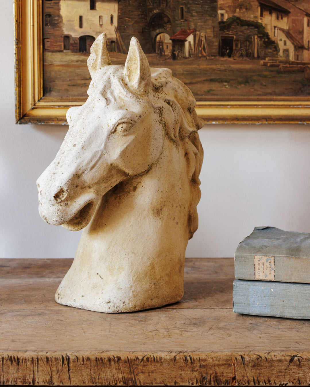 Light-colored horse head sculpture on a rustic wooden table, with a painting and books in the background.