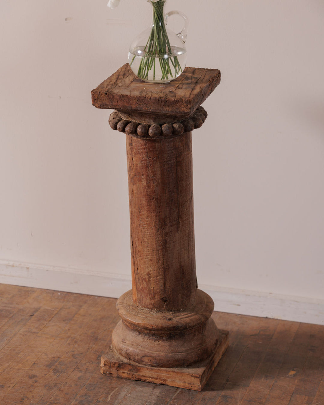 Rustic wooden pedestal with a small pitcher of green stems on top.