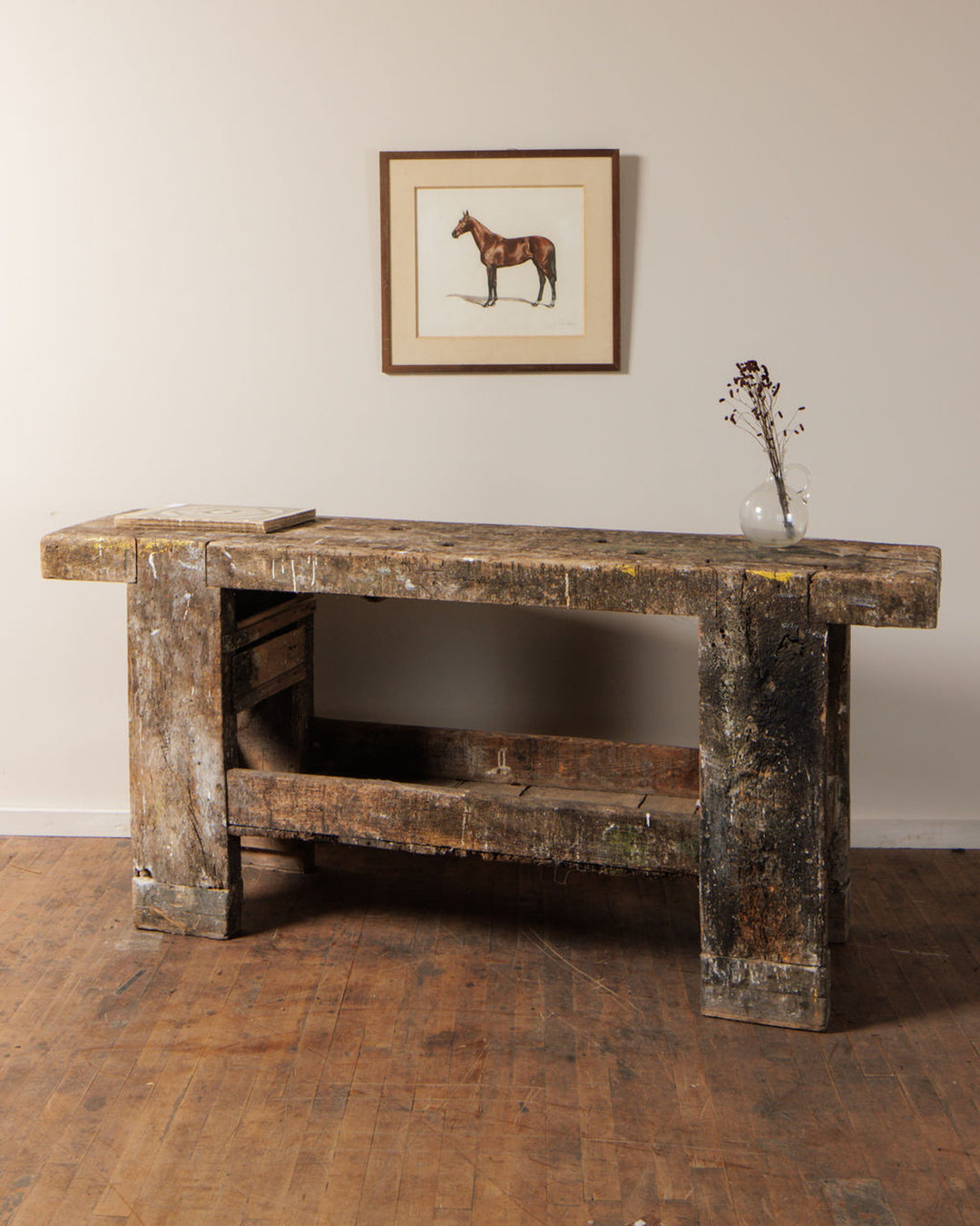 A distressed wooden workbench holds a book and dried flowers, below a framed horse painting on a light wall.