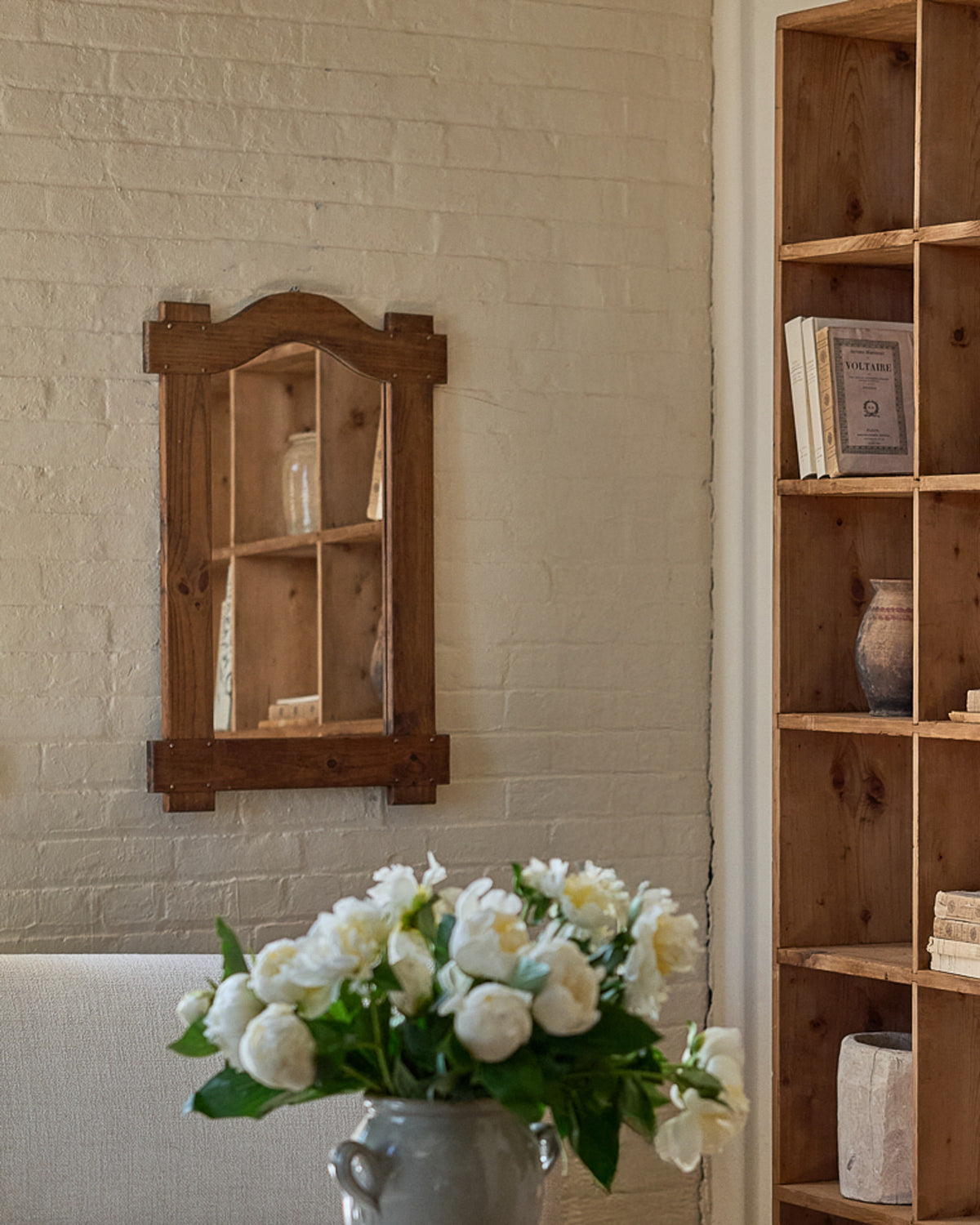 A rustic wooden mirror hangs on a light brick wall, with a vase of white peonies in the foreground and a wooden bookshelf on the right.