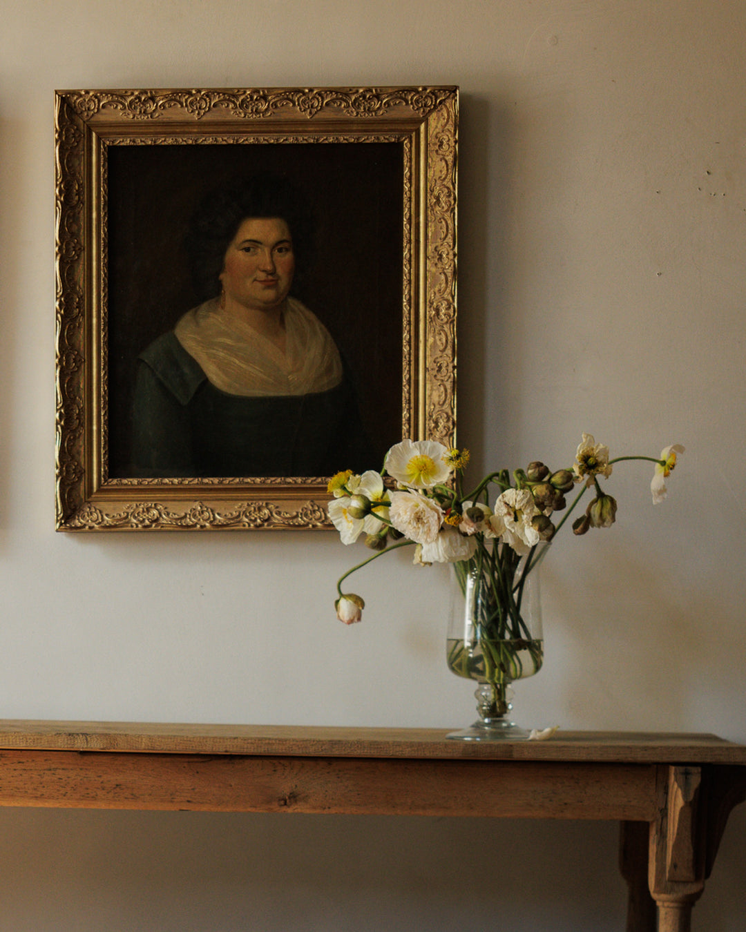 A gold-framed portrait of a woman hangs above a wooden table holding a vase of white and yellow flowers.