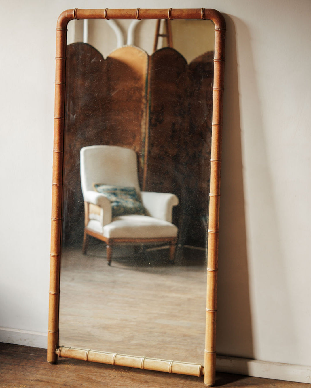 Tall mirror with a bamboo frame reflecting an armchair and interior.
