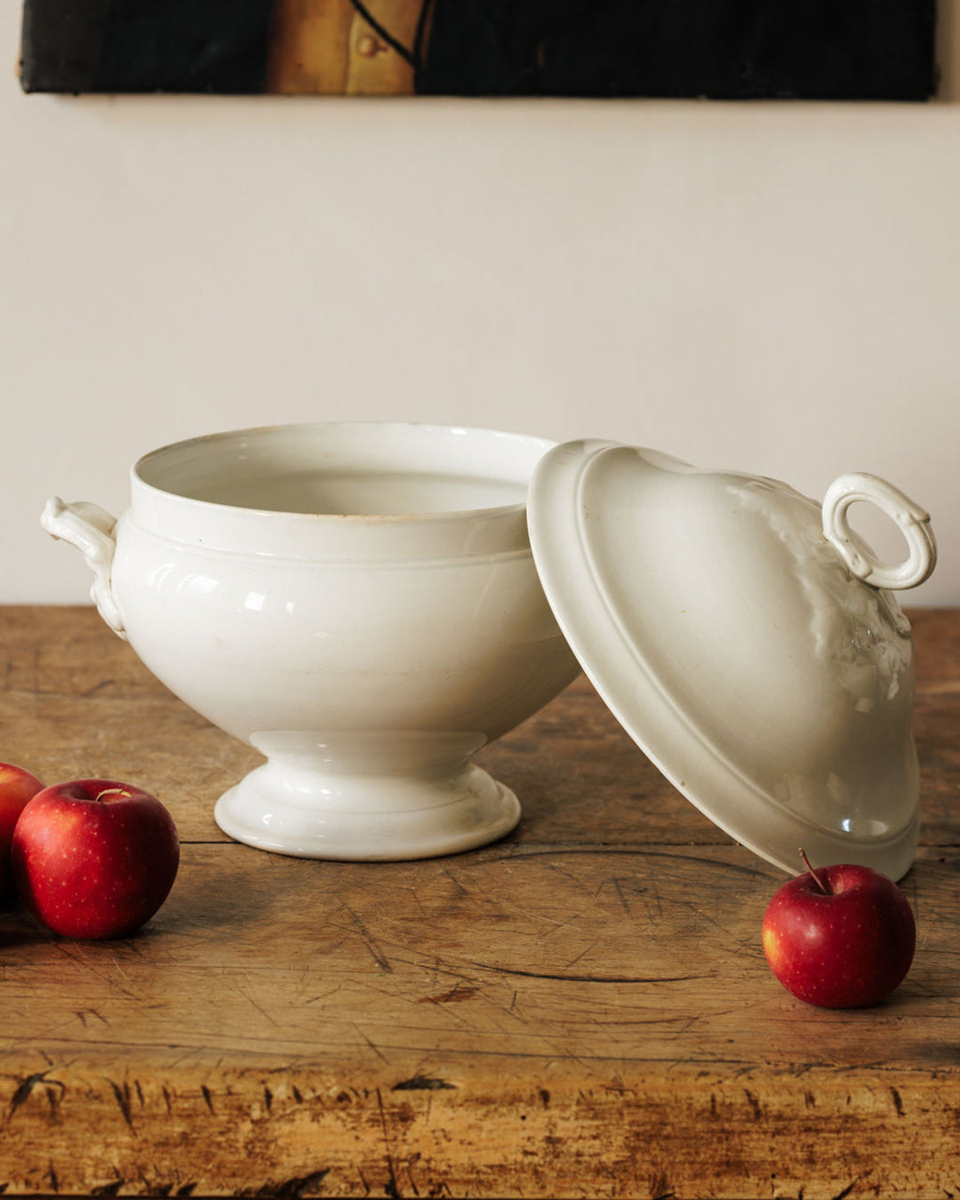 A white ceramic tureen with its lid leaning against it, on a wooden table with red apples.