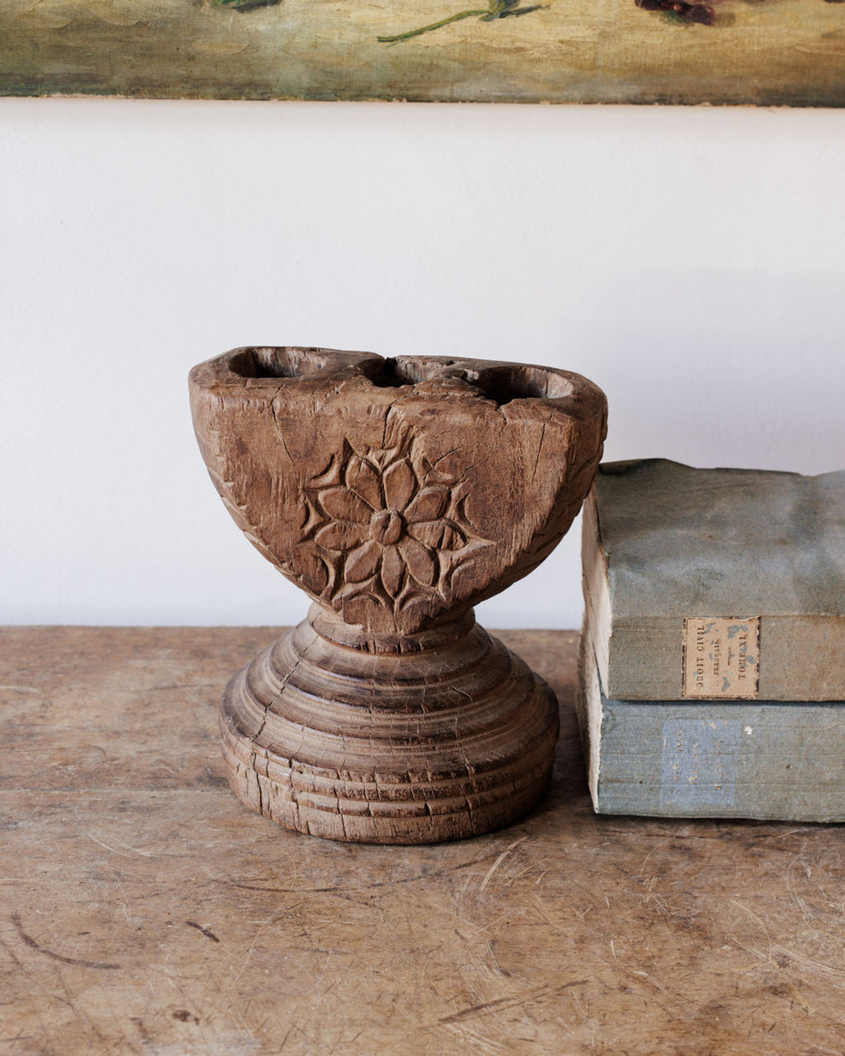 A rustic carved wooden holder with a floral pattern and three openings sits on a wooden table, next to a stack of old books.