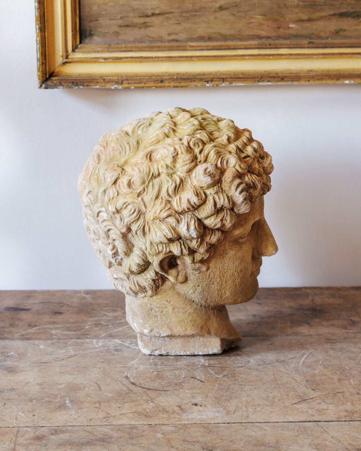 Profile view of a classical-style head sculpture with curly hair, on a wooden table beneath a framed painting.