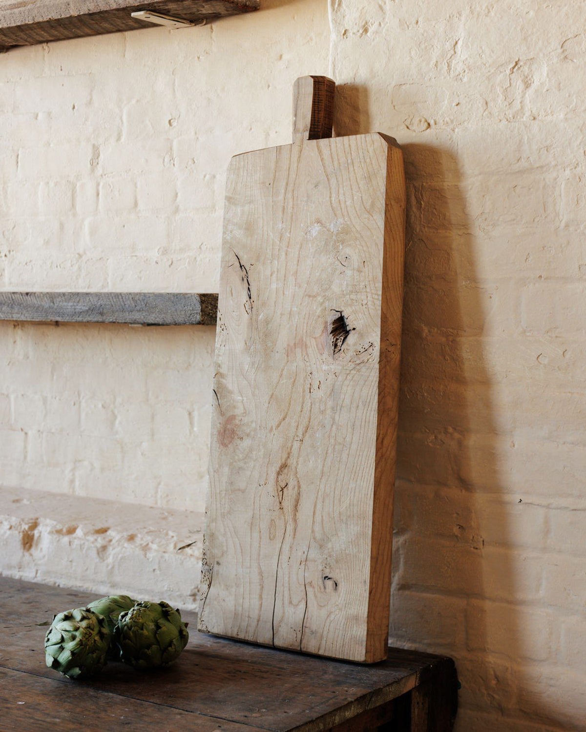 A large, rustic wooden cutting board leans against a white brick wall, with two artichokes on a wooden table.