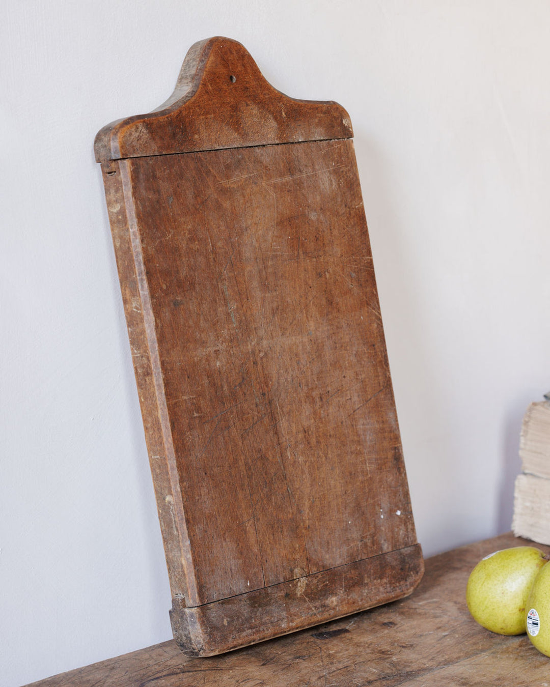 A rustic wooden breadboard with a curved handle top, leaning against a white wall.