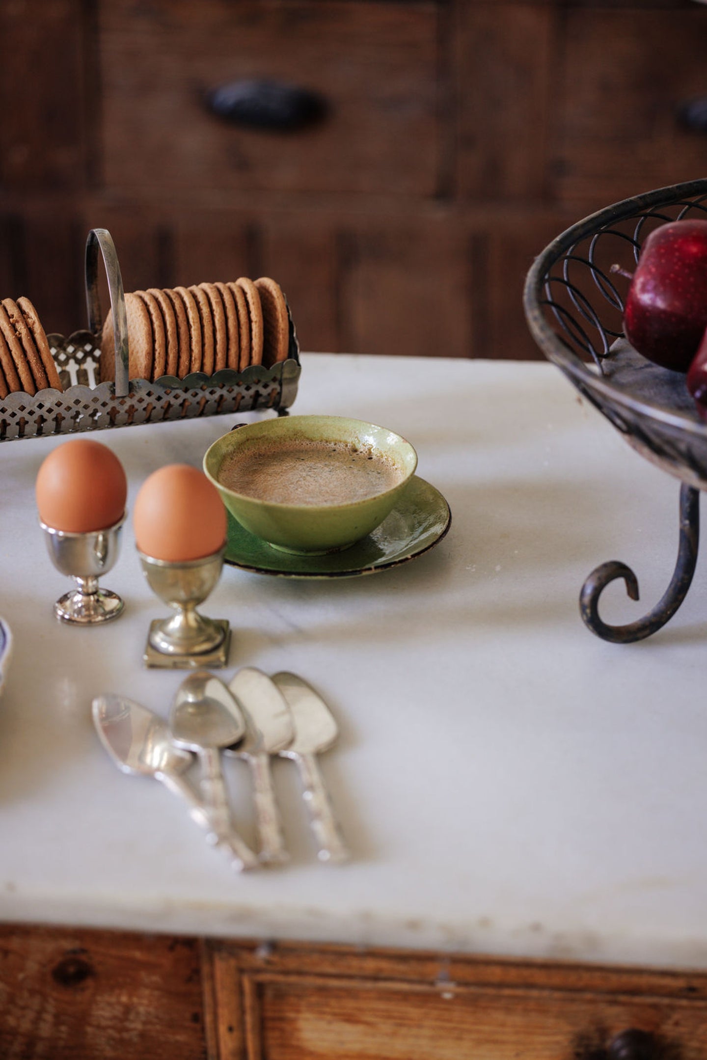 Breakfast items: a cup of coffee, two eggs in holders, biscuits, and apples on a marble counter.