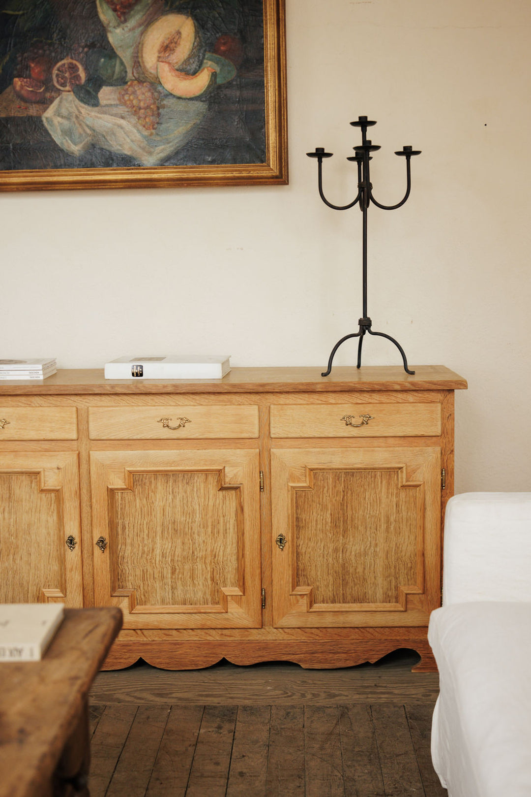 A wooden sideboard topped with a black candelabra and books, below a still-life painting, with a wooden floor and white furniture.