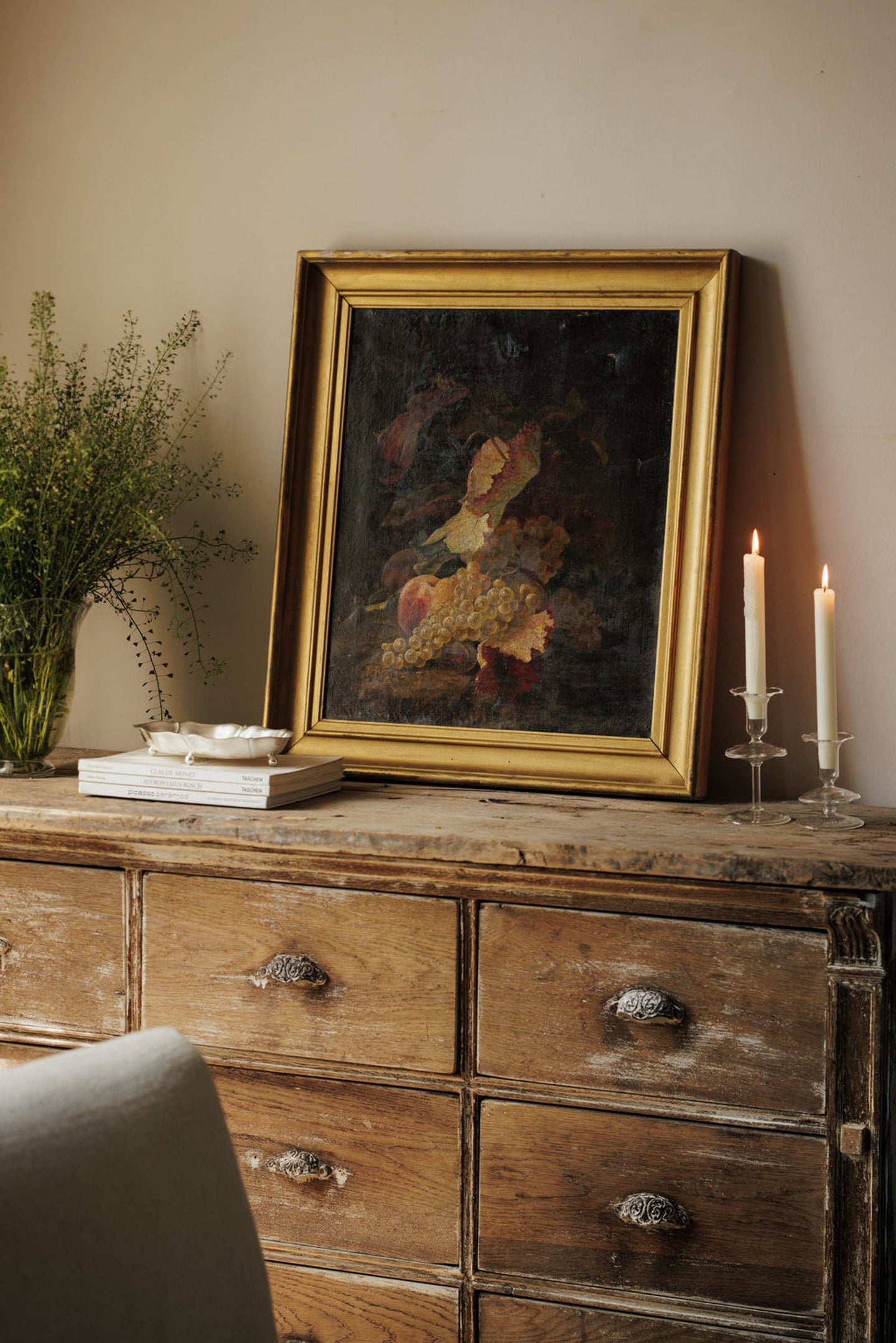 A rustic wooden dresser displaying a still life fruit painting, a vase of greenery, and two lit candles.
