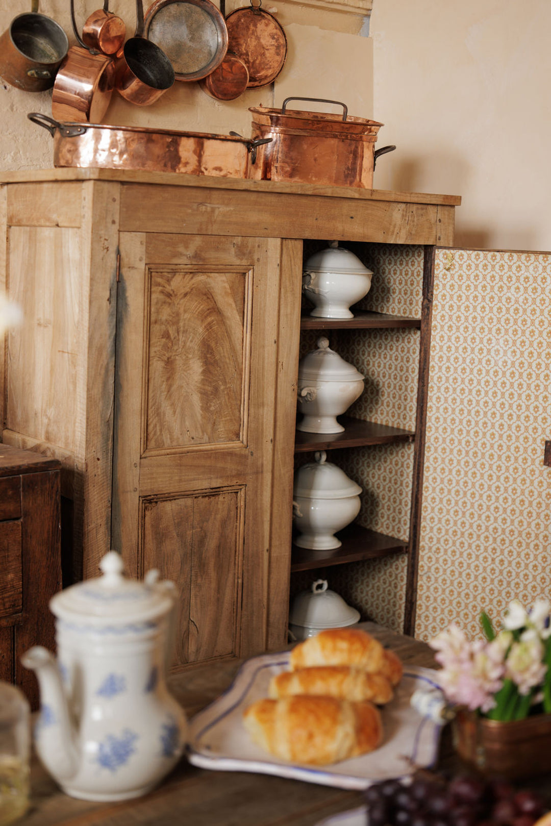 A wooden cabinet with an open door revealing white tureens, topped and surrounded by copper cookware, next to a table with croissants and a teapot.
