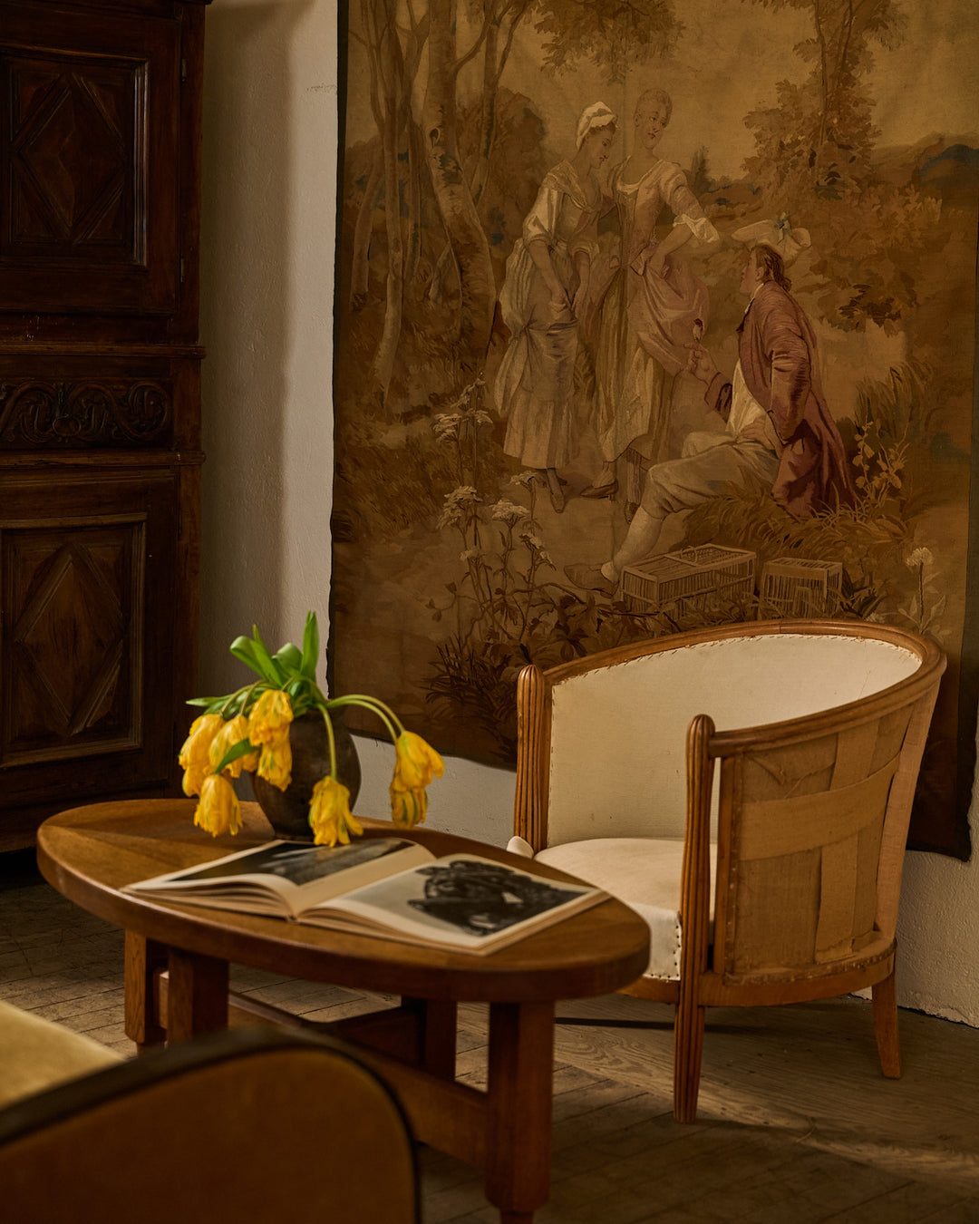 A vintage room with a large tapestry depicting figures, an oval wooden table with yellow tulips and an open photo book, and an upholstered armchair.