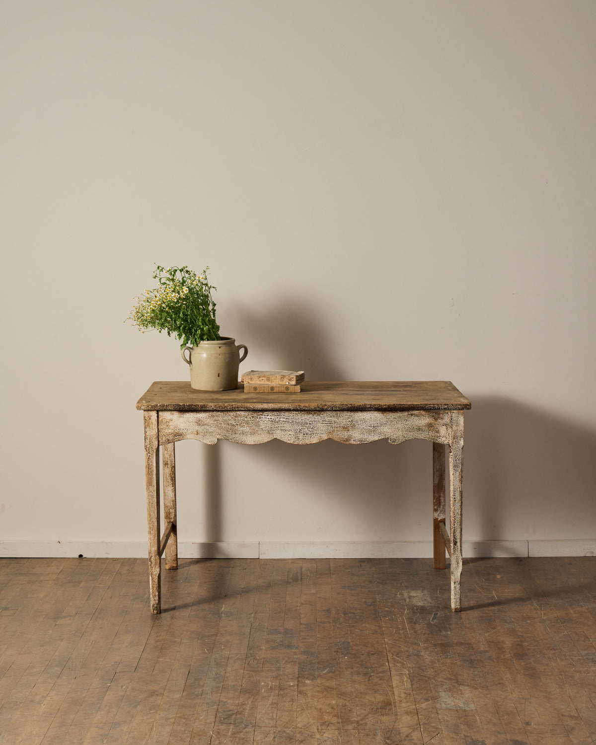 Distressed wooden console table with a scalloped apron, chipped white paint, a vase of wildflowers, and a small wooden box.