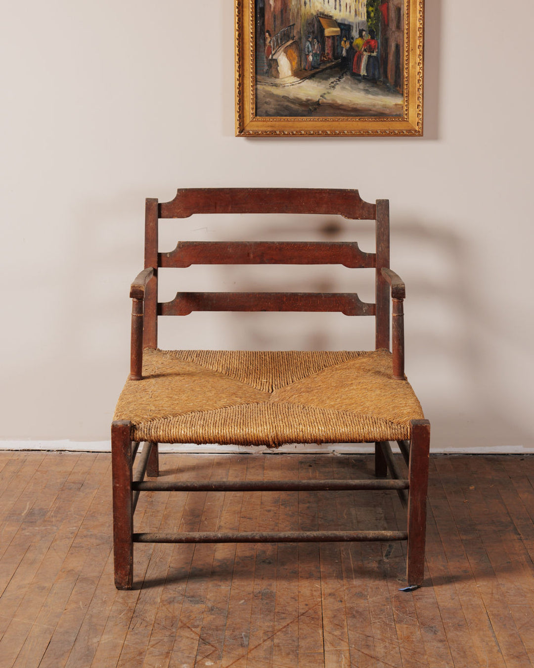 A rustic dark wood ladder-back chair with a rush seat sits on a wooden floor beneath a framed painting.