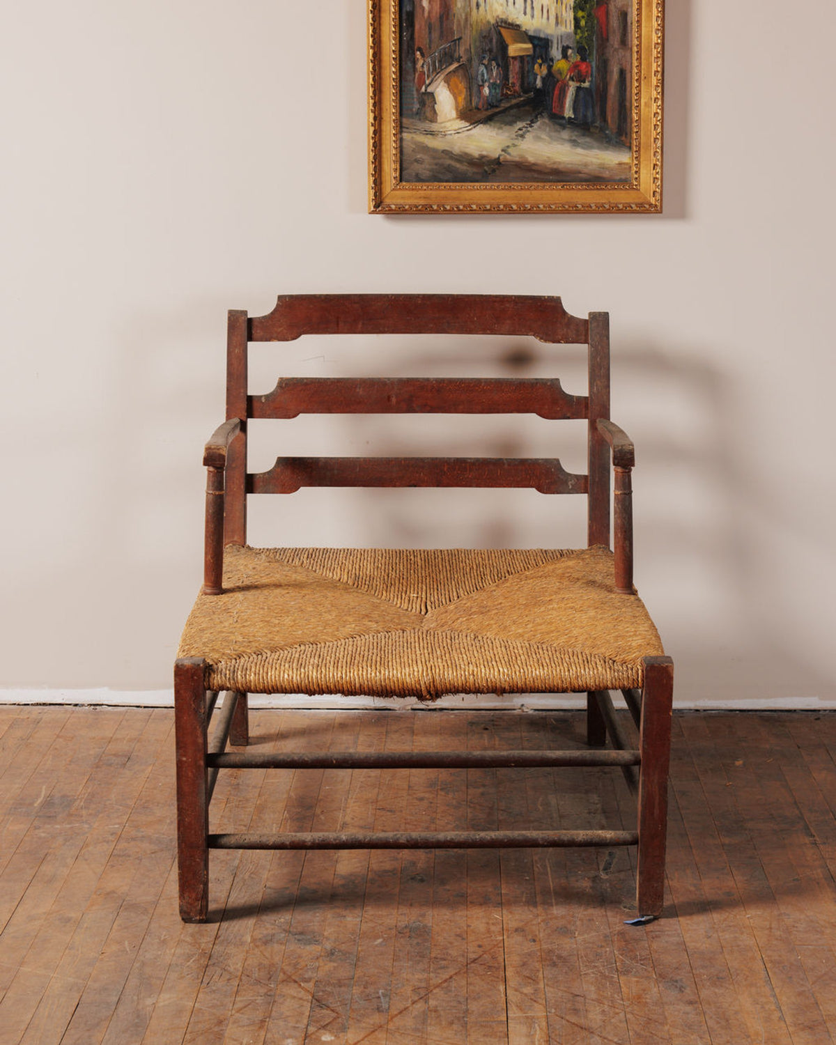 A rustic dark wood ladder-back chair with a rush seat sits on a wooden floor beneath a framed painting.