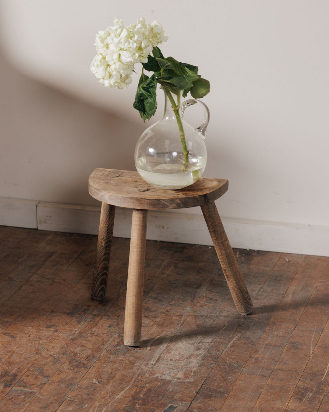 A glass pitcher with white hydrangeas on a rustic half-moon wooden stool.