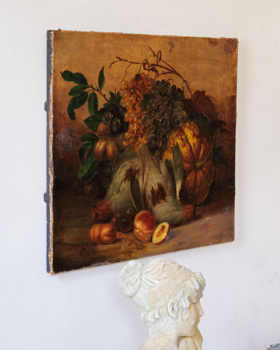 A classical still life painting of fruit and fowl, with a partial stone bust in the foreground.