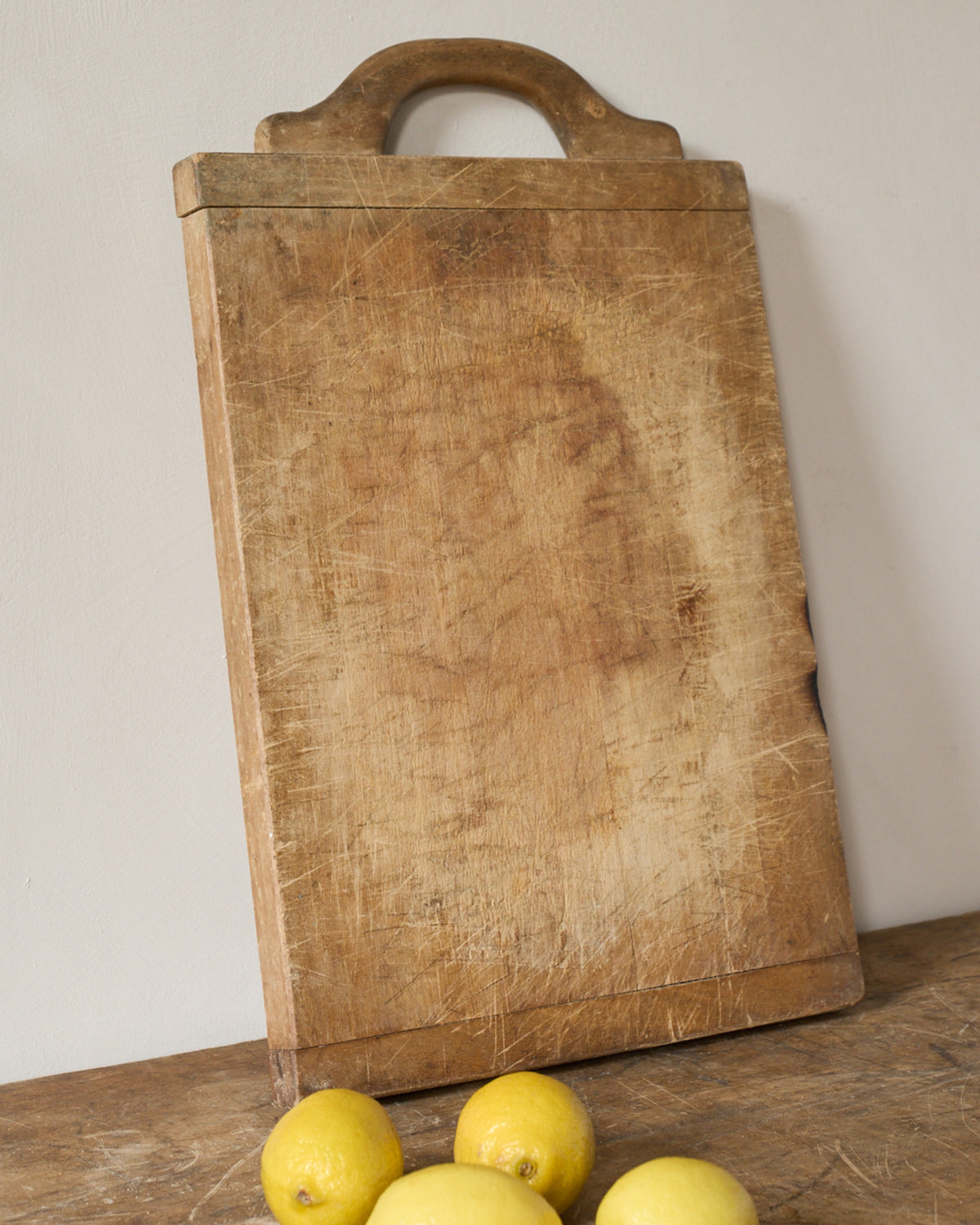 A well-worn wooden cutting board with a handle leans against a wall, with several lemons on a wooden surface below.