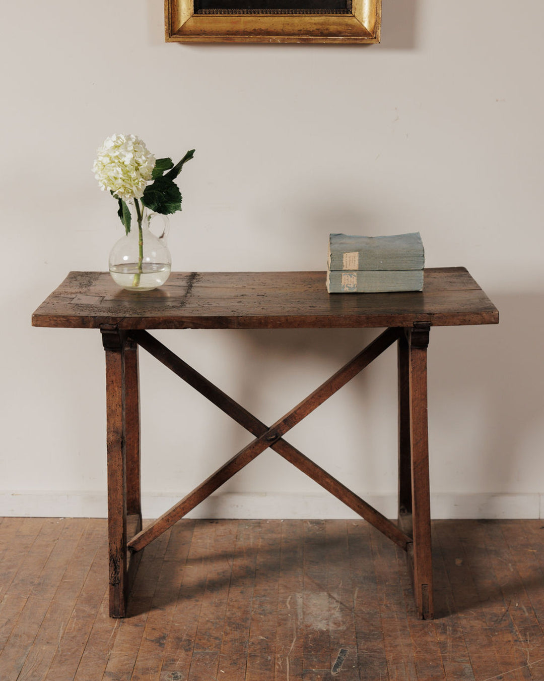A rustic wooden table with X-braced legs, holding a vase of white hydrangeas and two light blue books.