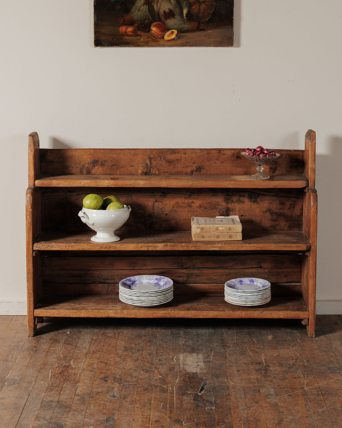 A rustic three-tier wooden shelf displaying fruit, books, and patterned plates, with a painting above.