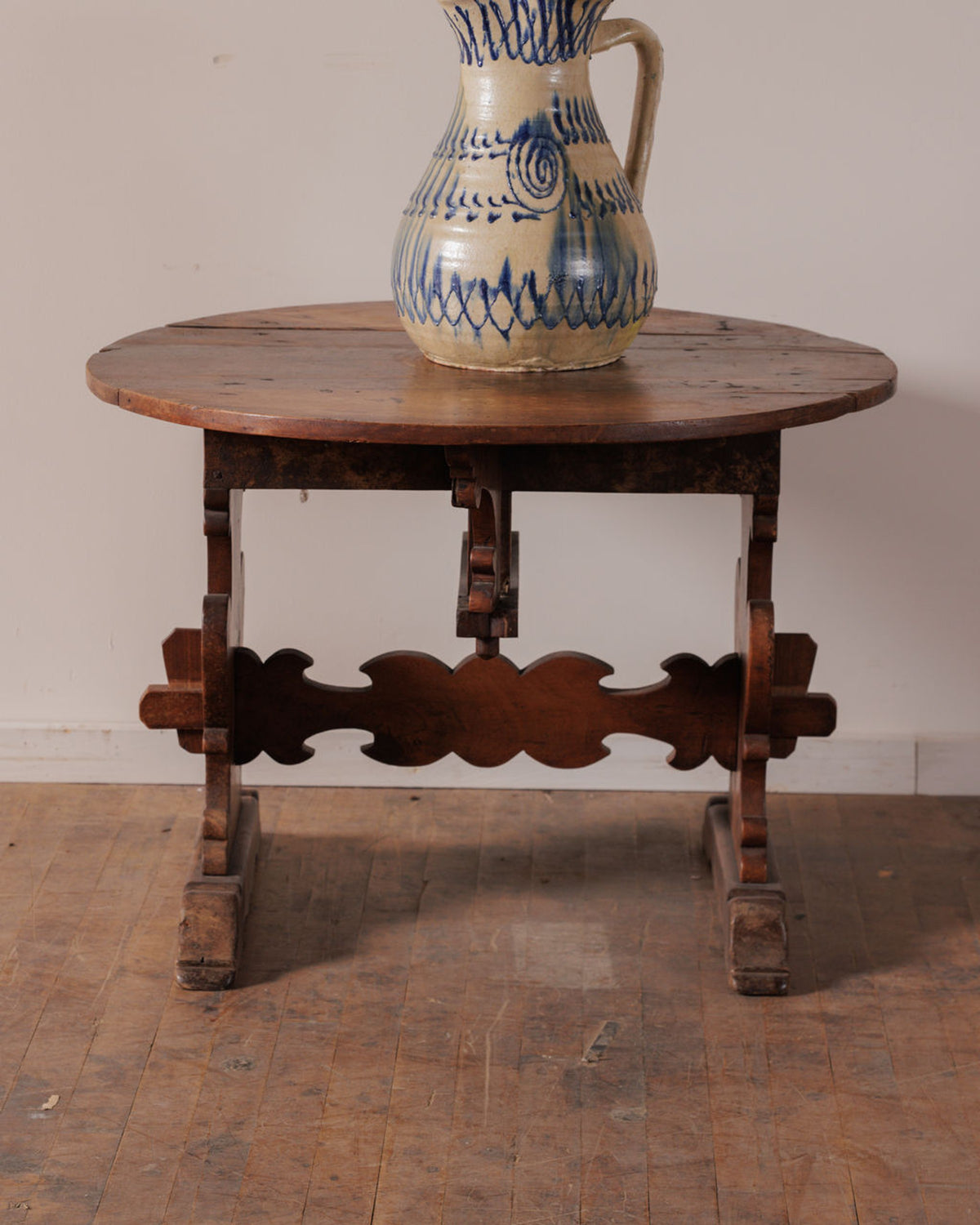 A cream-colored, blue-patterned ceramic pitcher sits on a round, rustic wooden table with an ornate carved base.