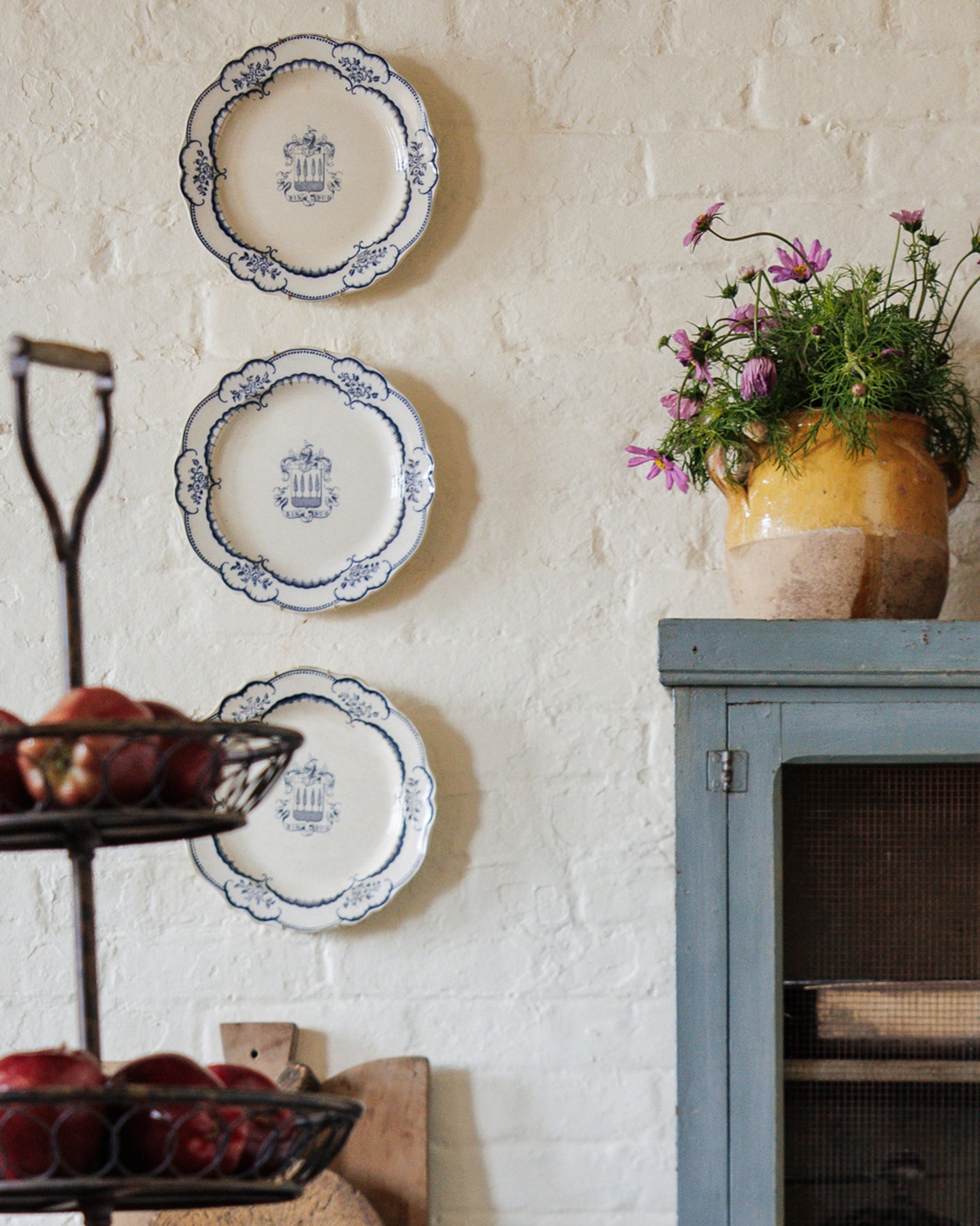 Three decorative blue and white plates on a white brick wall, with a pot of pink flowers on a blue cabinet and apples in a tiered basket.