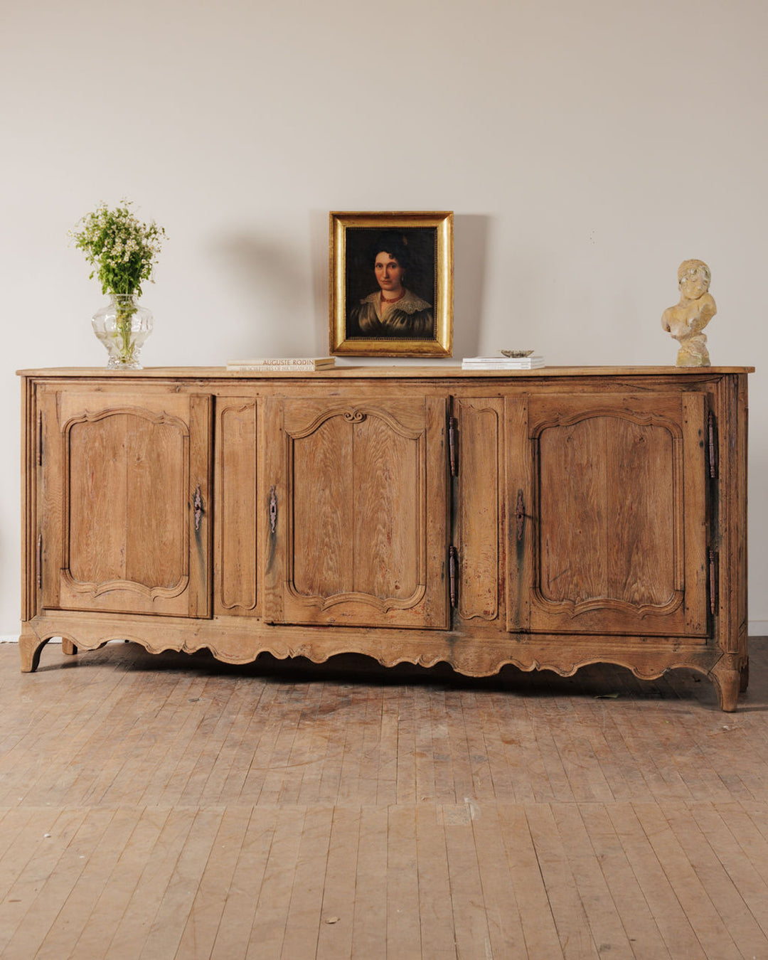 A light wood four-door antique buffet with a vase of flowers, a framed portrait, and a bust on top.