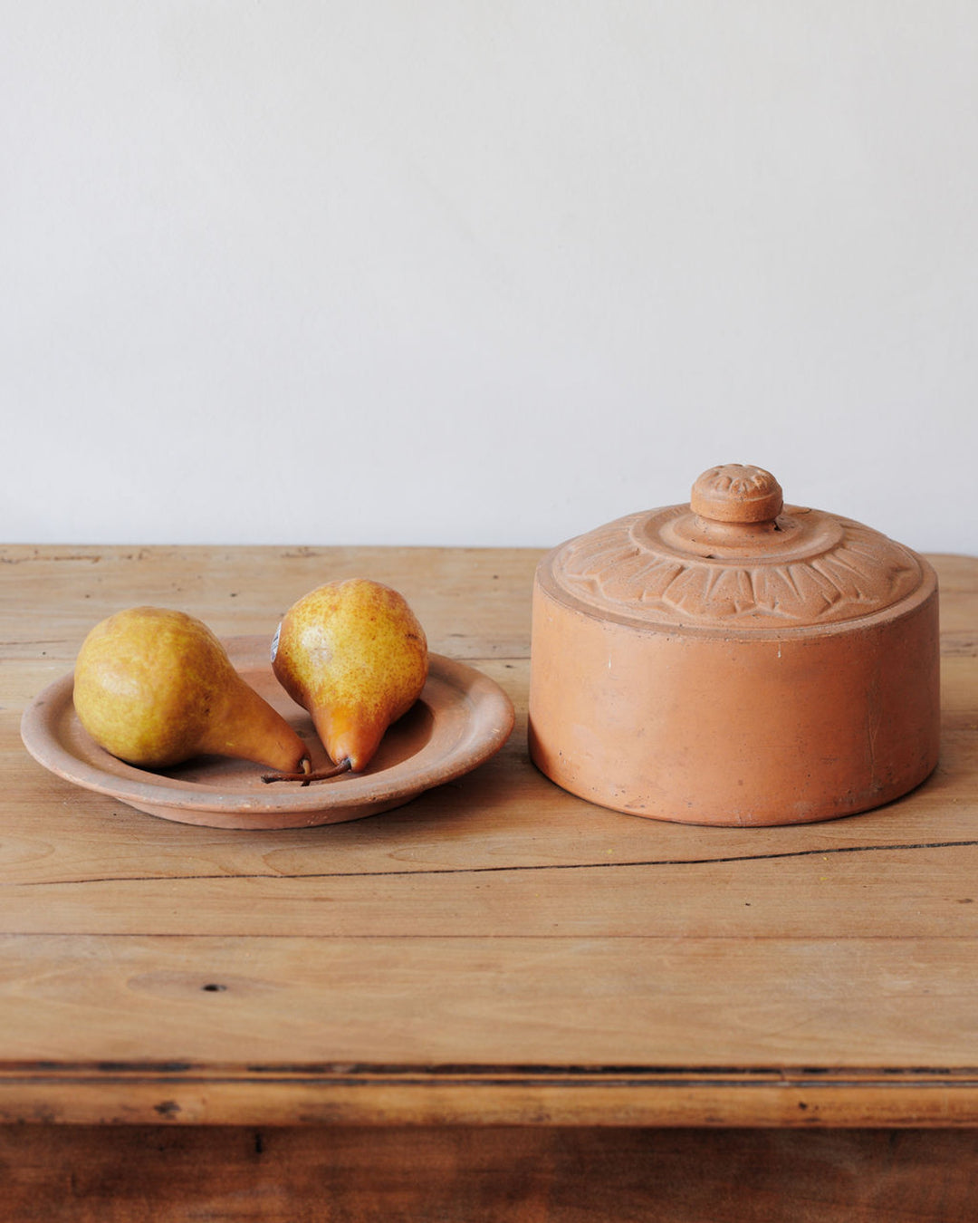 Two pears on a terracotta plate next to a lidded terracotta pot on a wooden table.