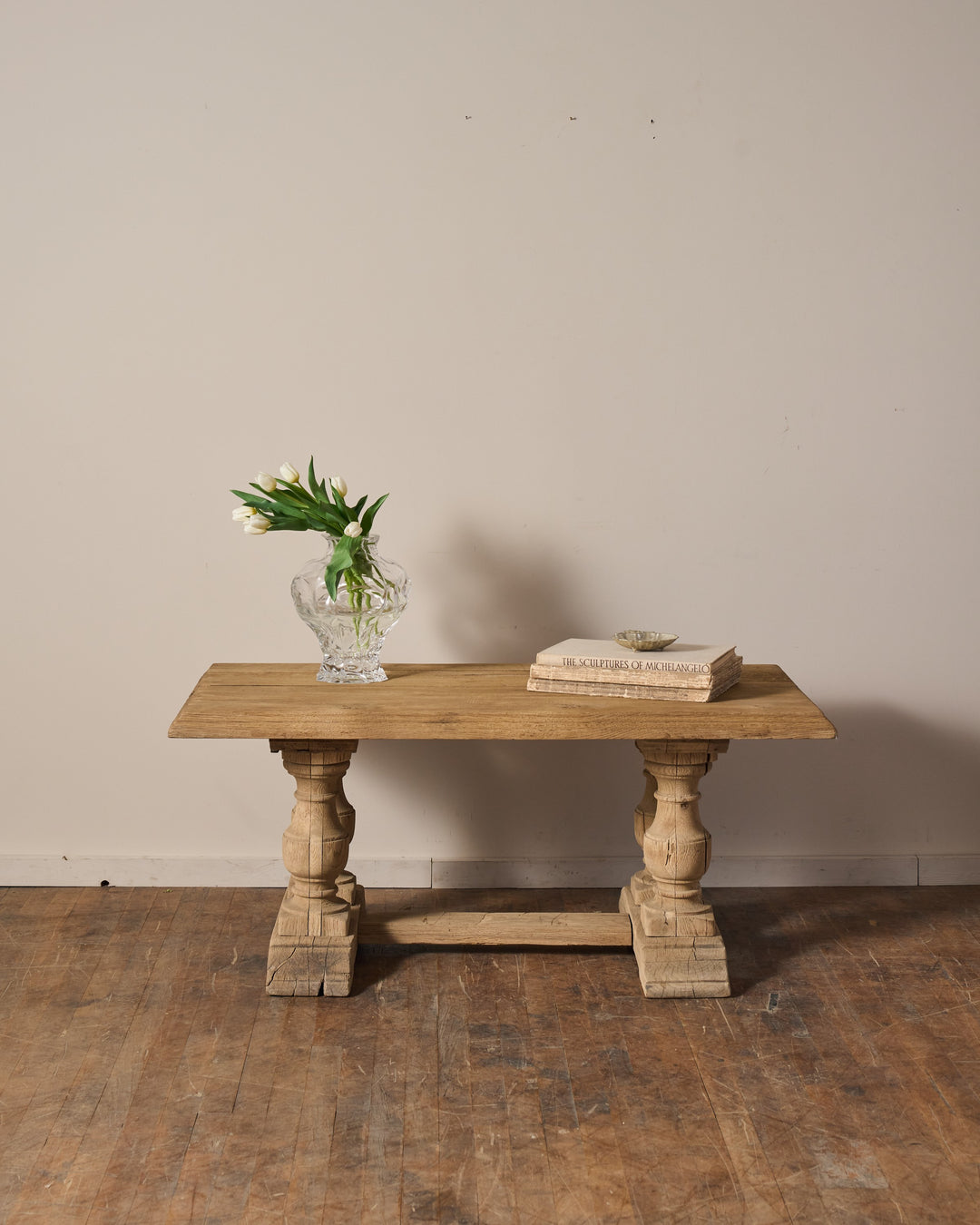 A rustic light wood console table with carved legs, holding a vase of white tulips and stacked books.