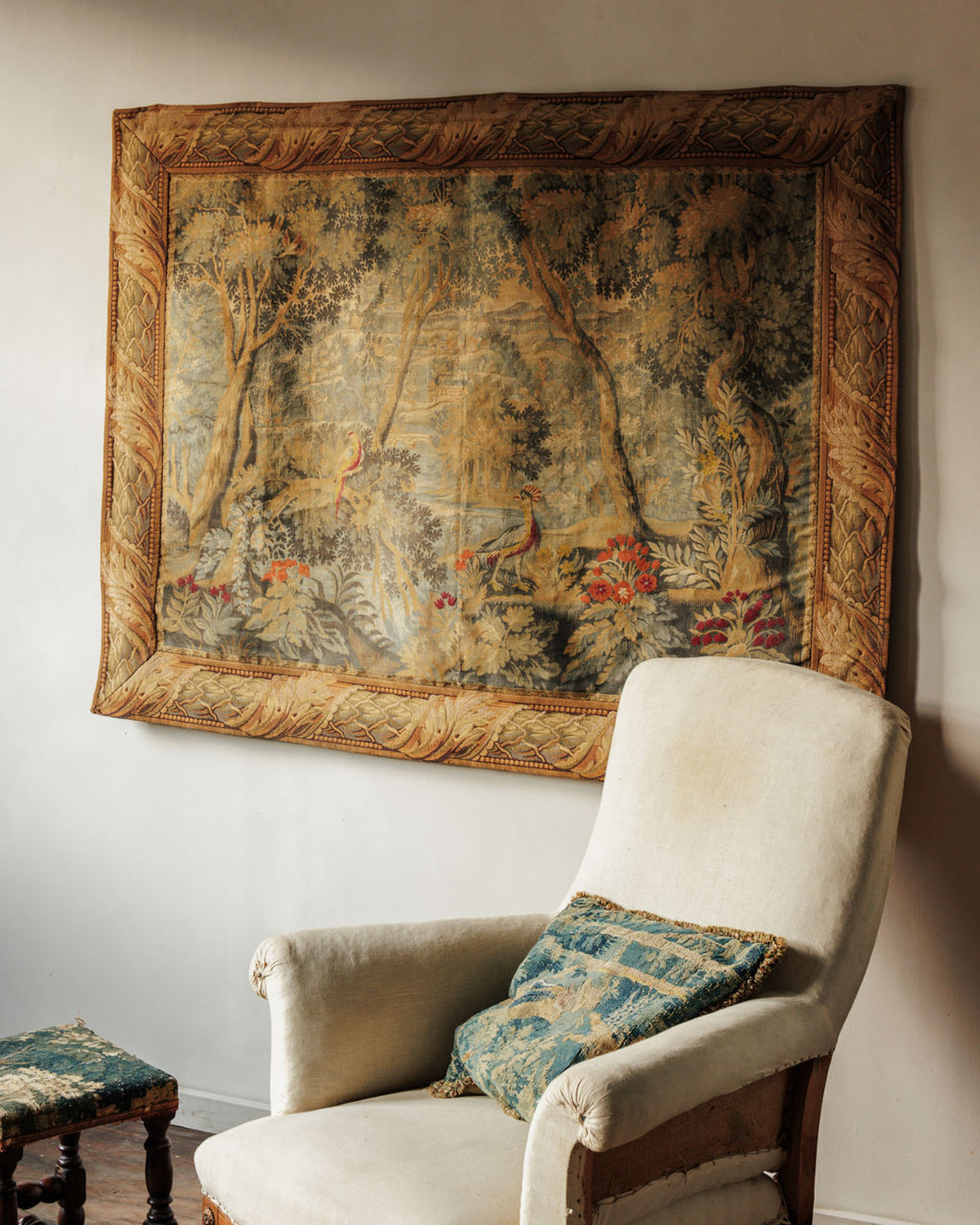 An antique tapestry depicting a woodland scene with birds hangs above a distressed cream armchair.