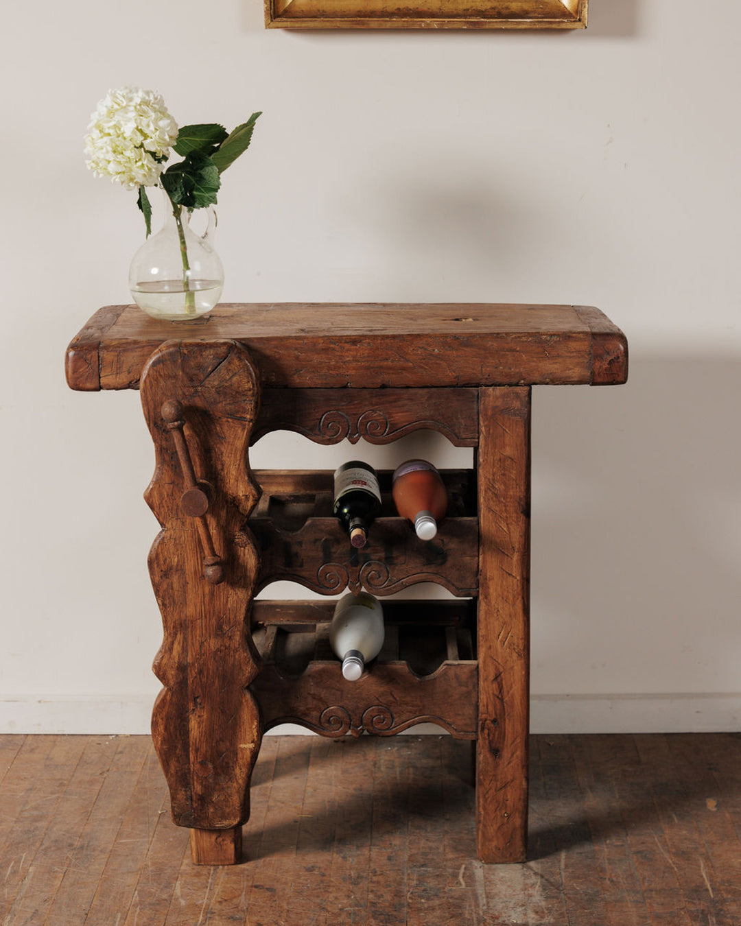 A rustic wooden wine rack with a vase of white hydrangeas on top and three wine bottles stored within.