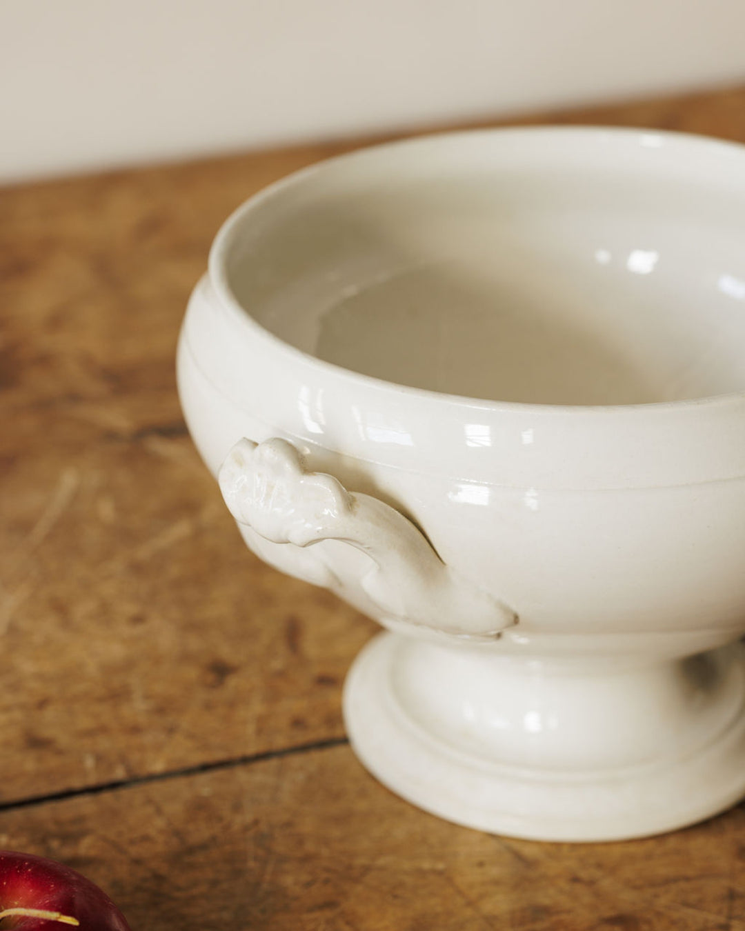White ceramic tureen with hand-shaped handles and a pedestal base on a wooden table.
