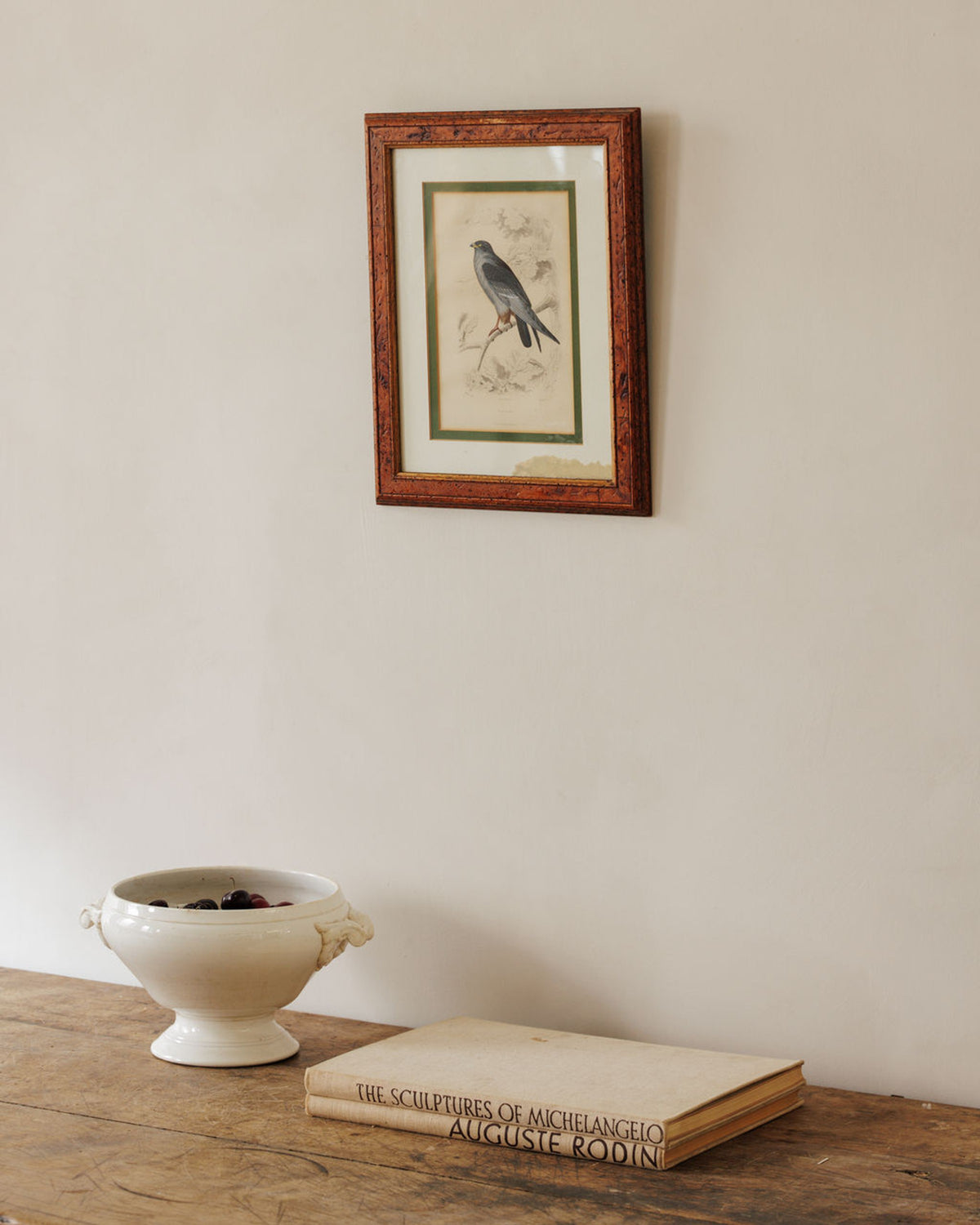 A framed bird illustration hangs on a light wall above a wooden table holding a white bowl of berries and a book titled "The Sculptures of Michelangelo Auguste Rodin."
