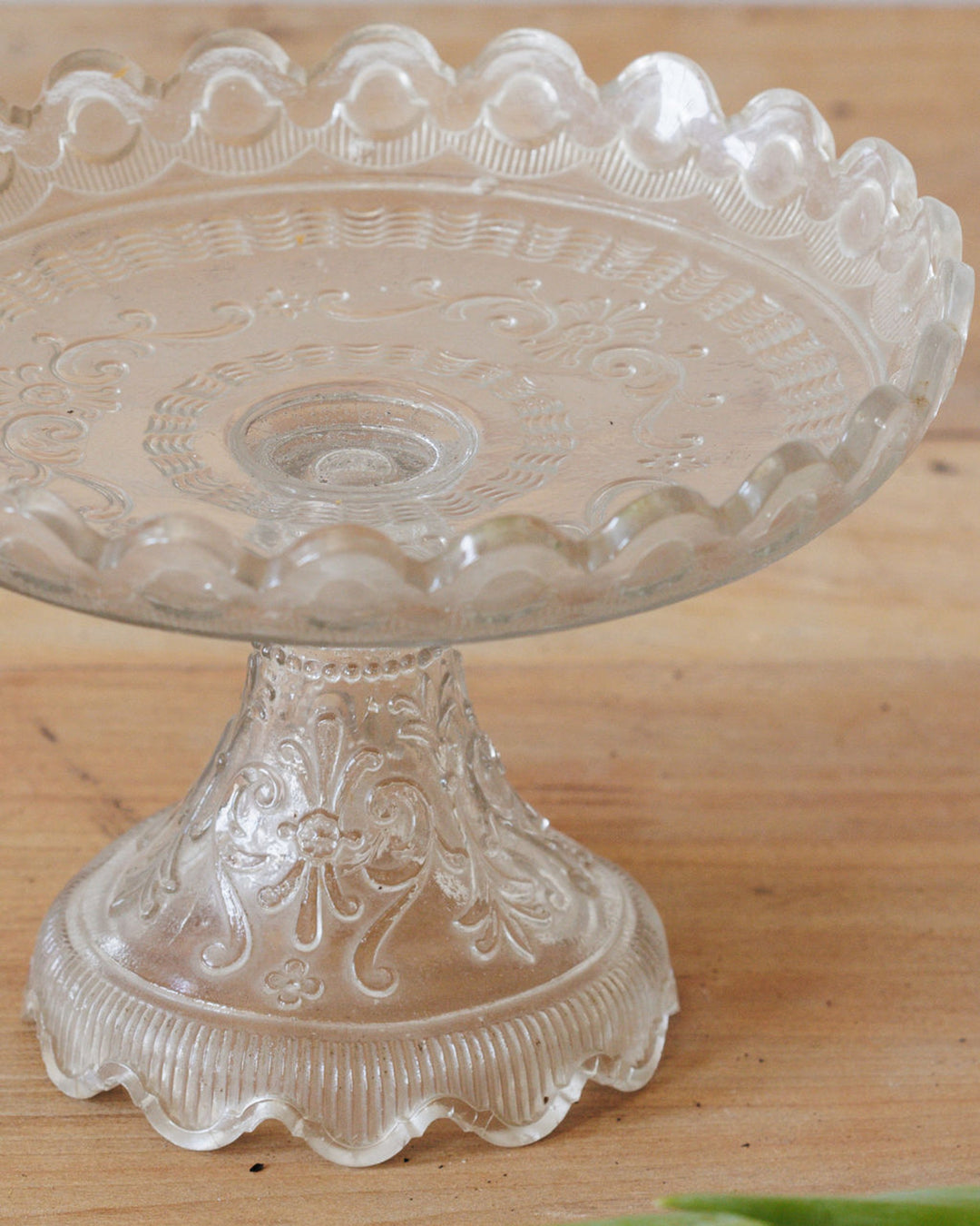 A clear glass cake stand with ornate embossed patterns and a scalloped edge.