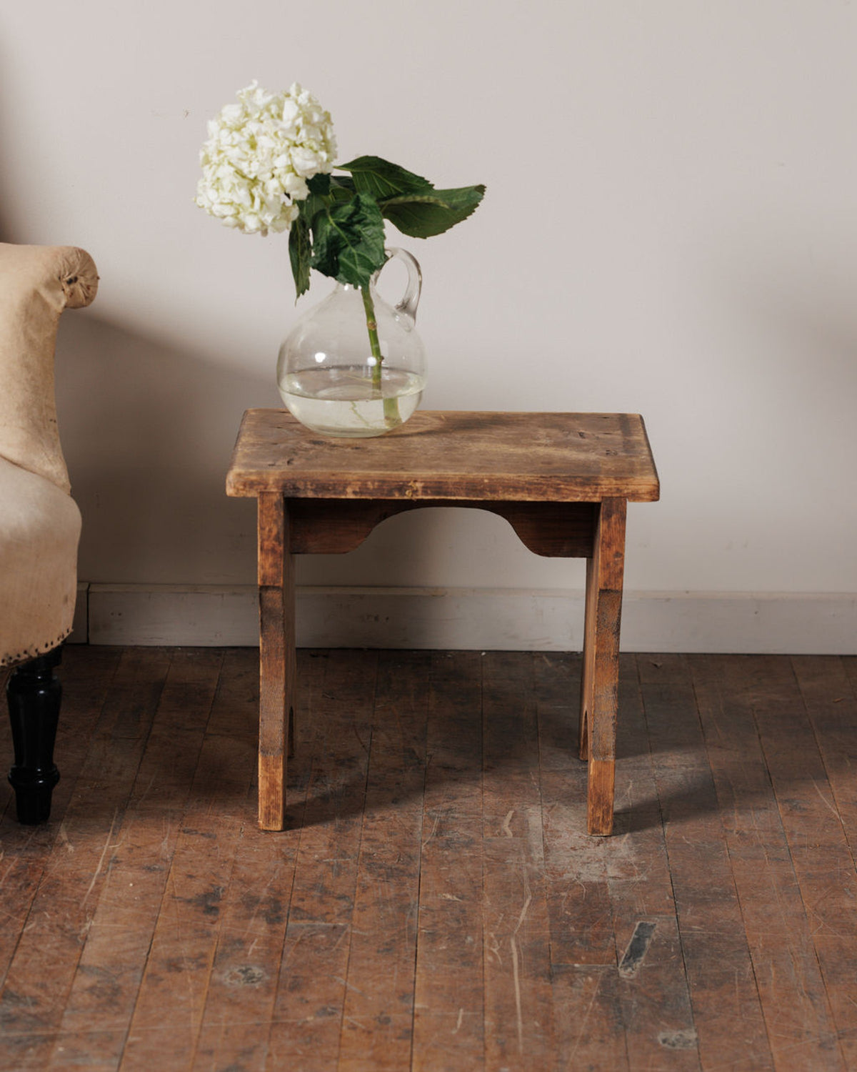 White hydrangea in a clear pitcher on a rustic wooden table next to a beige chair.