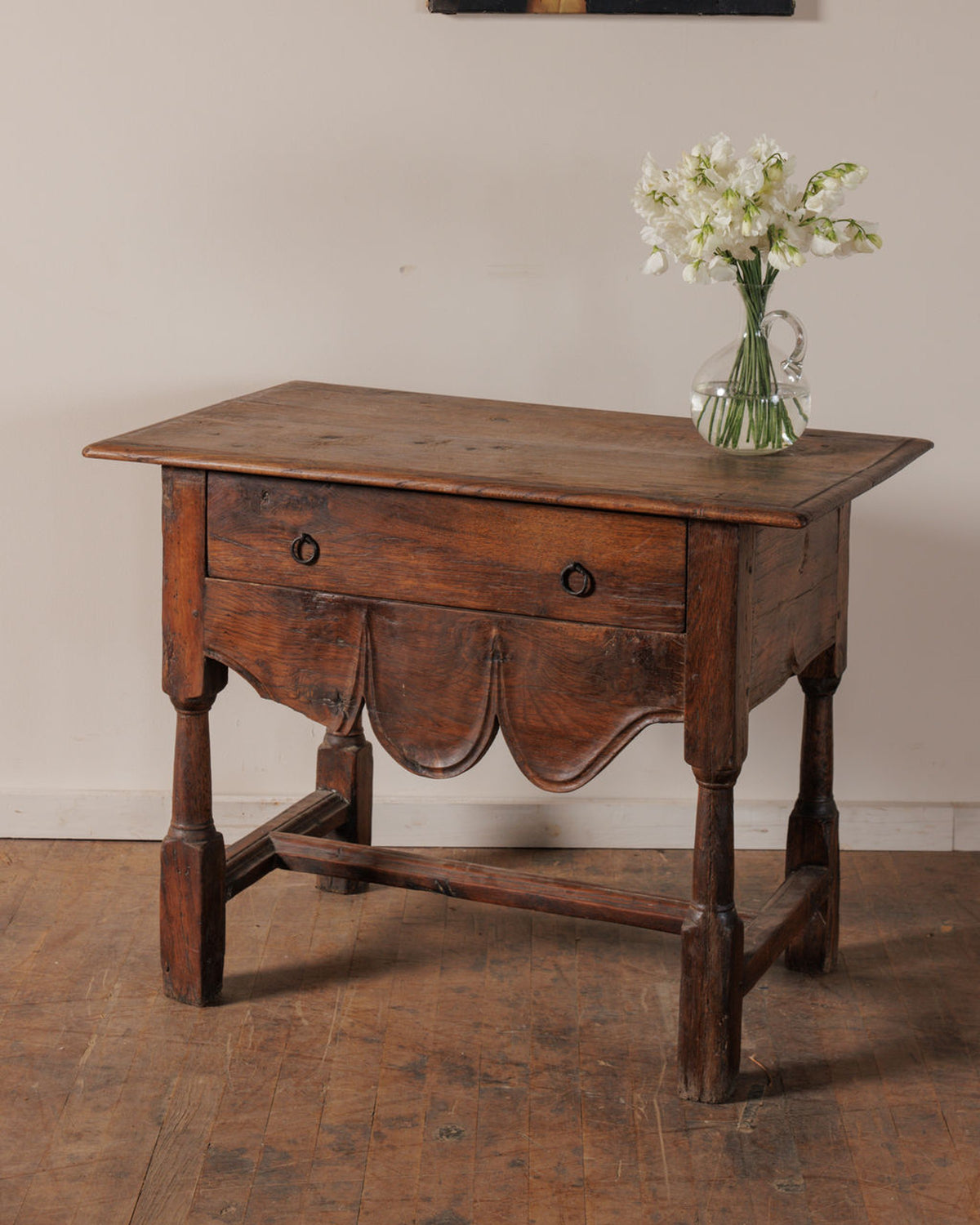 Antique rustic wooden table with a single drawer, scalloped apron, and a vase of white flowers.