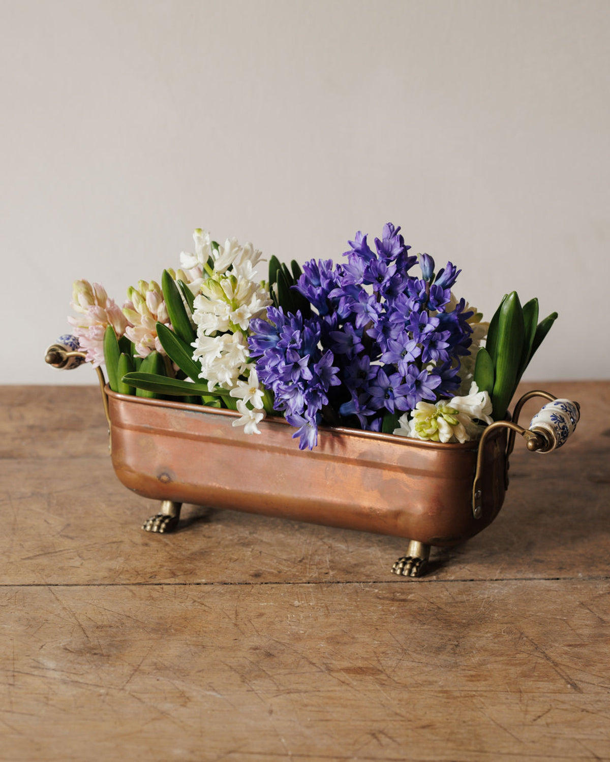 Miniature copper bathtub planter with pink, white, and purple hyacinths on a wooden surface.