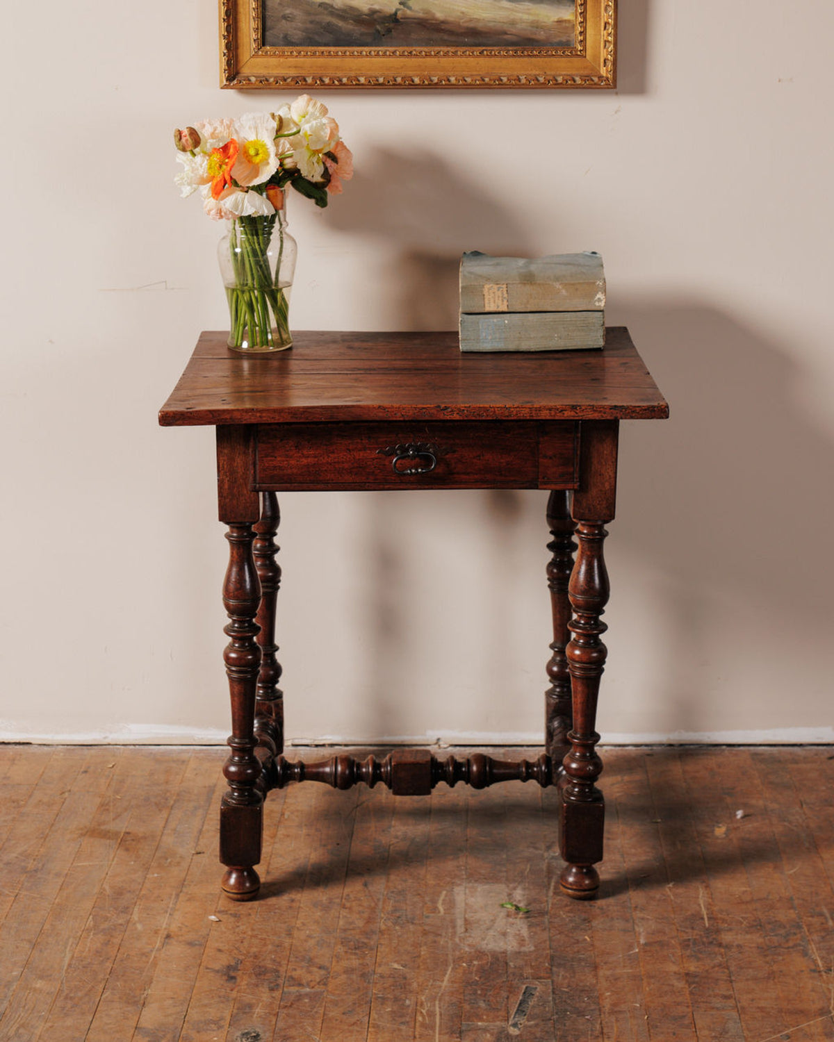 An antique dark wood table with turned legs, a single drawer, topped with a vase of colorful flowers and two light blue books.