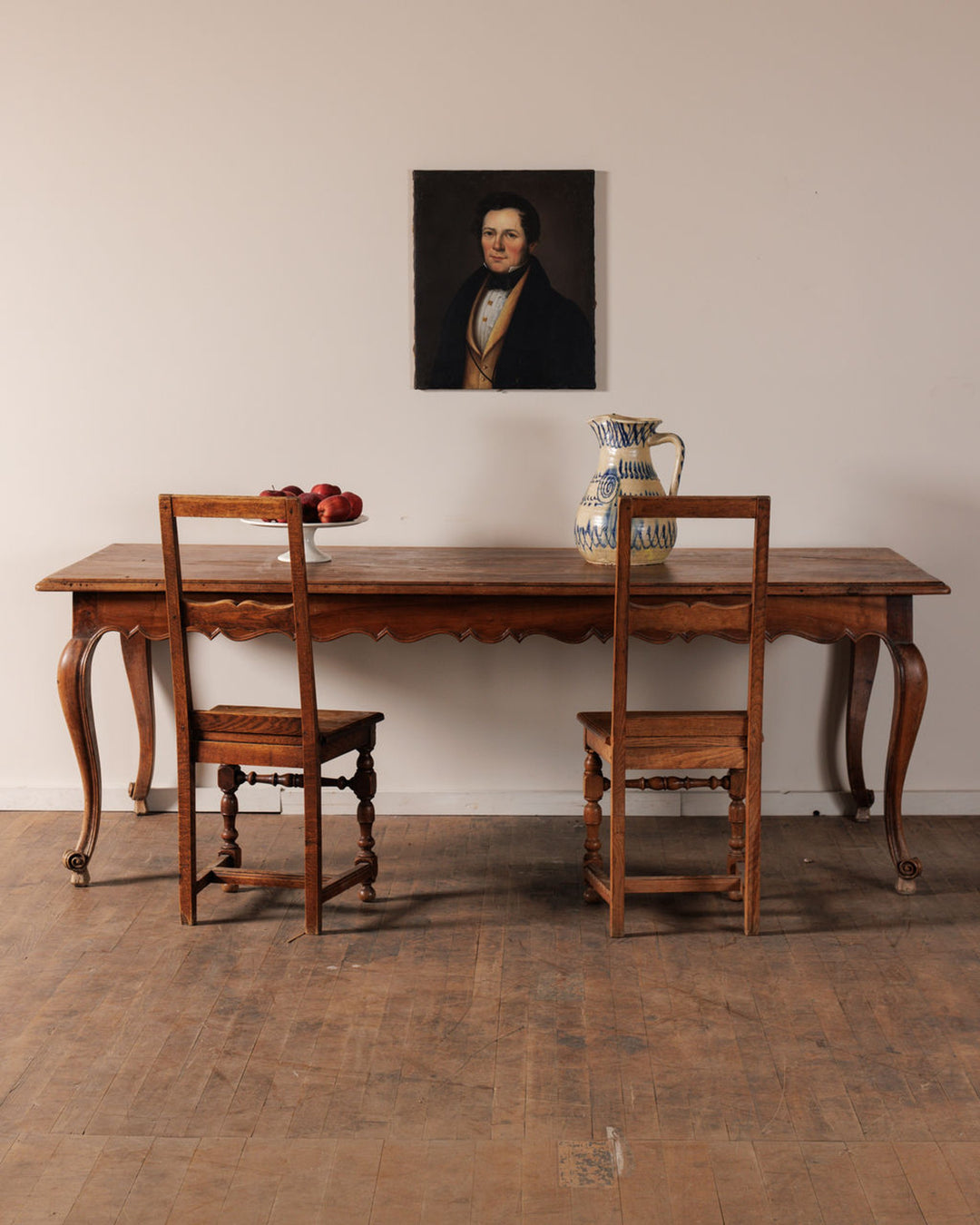Antique wooden dining table with two chairs, a bowl of apples, and a patterned pitcher, beneath a portrait painting on a light wall.