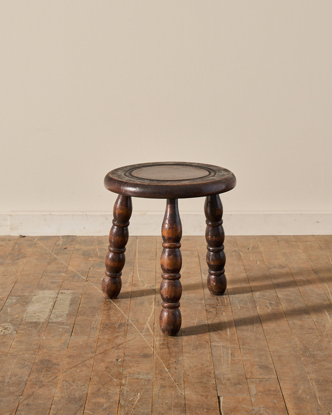 A dark brown wooden stool with a circular top and three turned legs sits on a worn wooden floor.