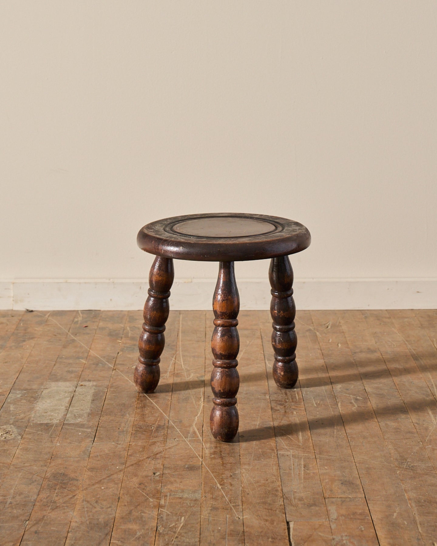 A dark brown wooden stool with a circular top and three turned legs sits on a worn wooden floor.
