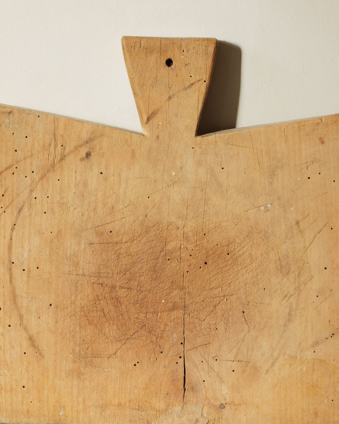 Old, light wood cutting board with scratches, small dark holes, and a flared handle.