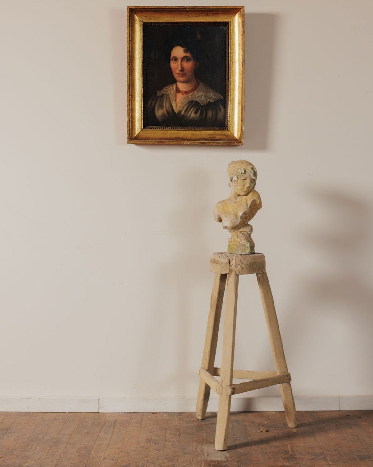 A framed portrait painting of a woman hangs above a bust sculpture displayed on a tall wooden stool.