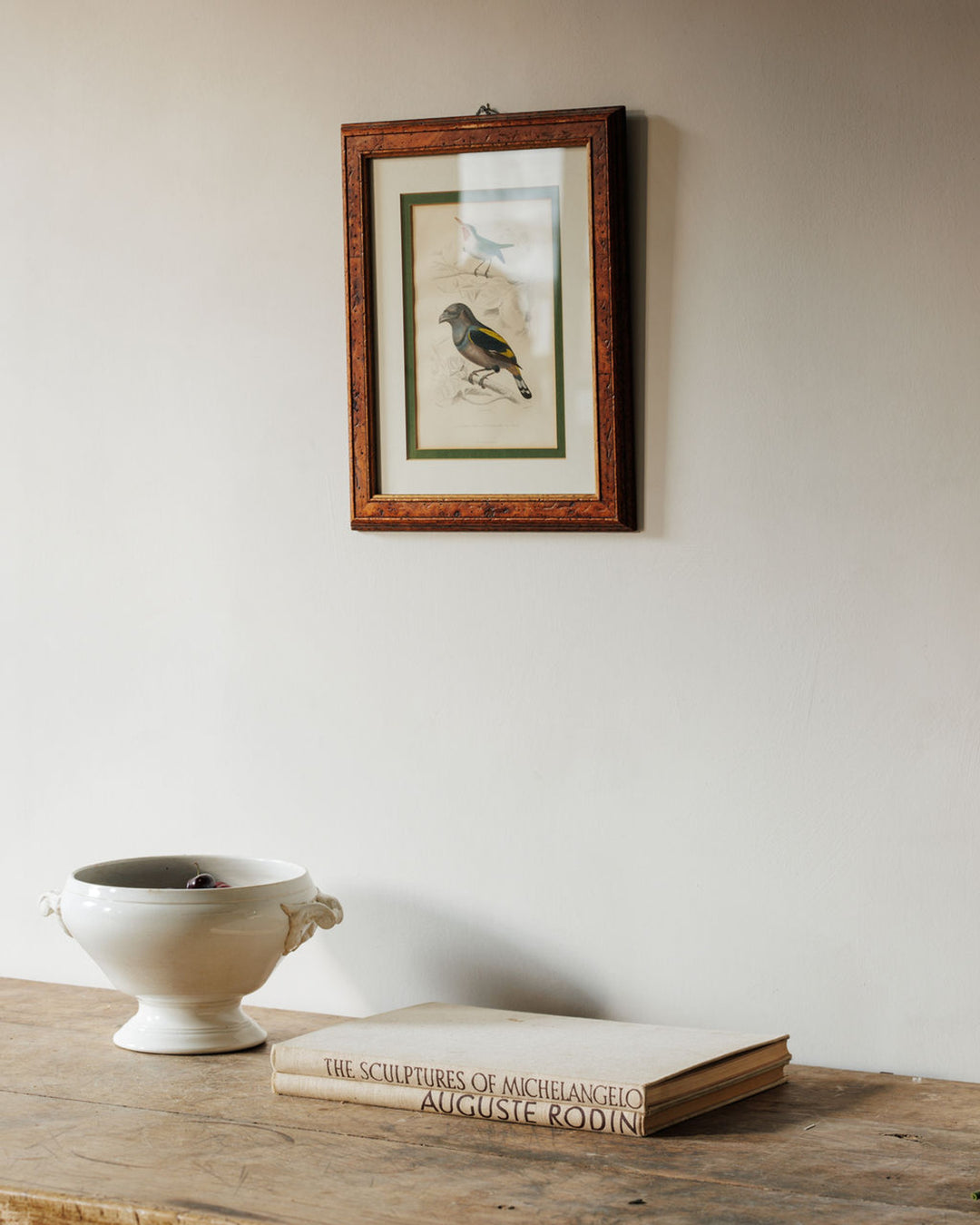 A framed bird illustration hangs above a white bowl and a book on a rustic wooden table.