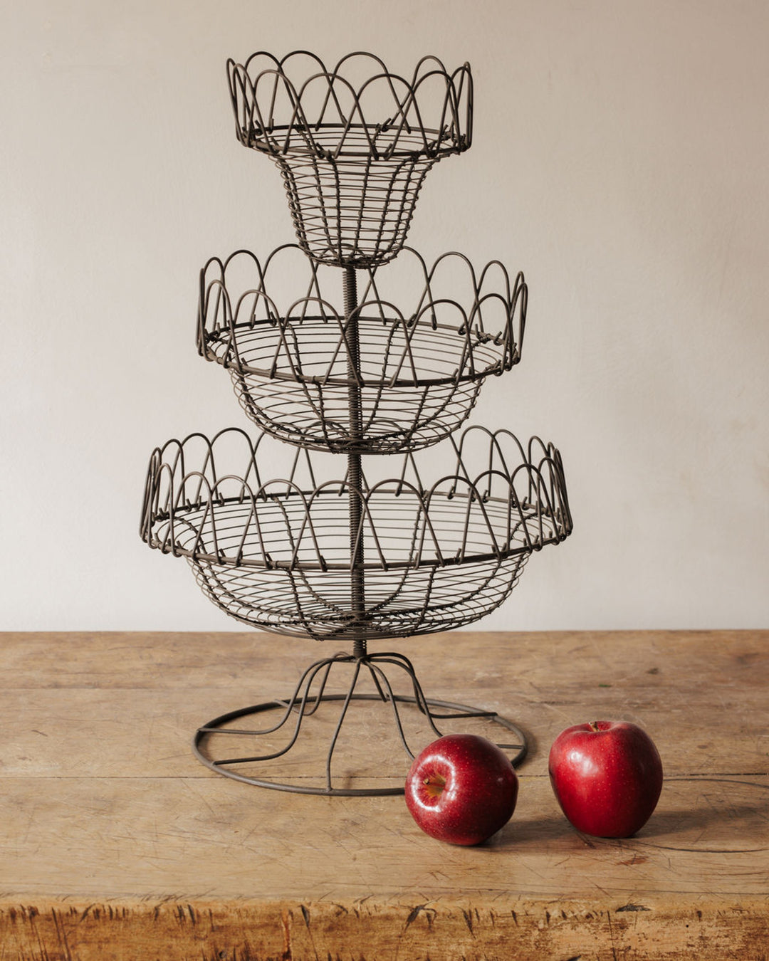 A three-tiered dark metal wire fruit basket on a wooden table with two red apples beside it.