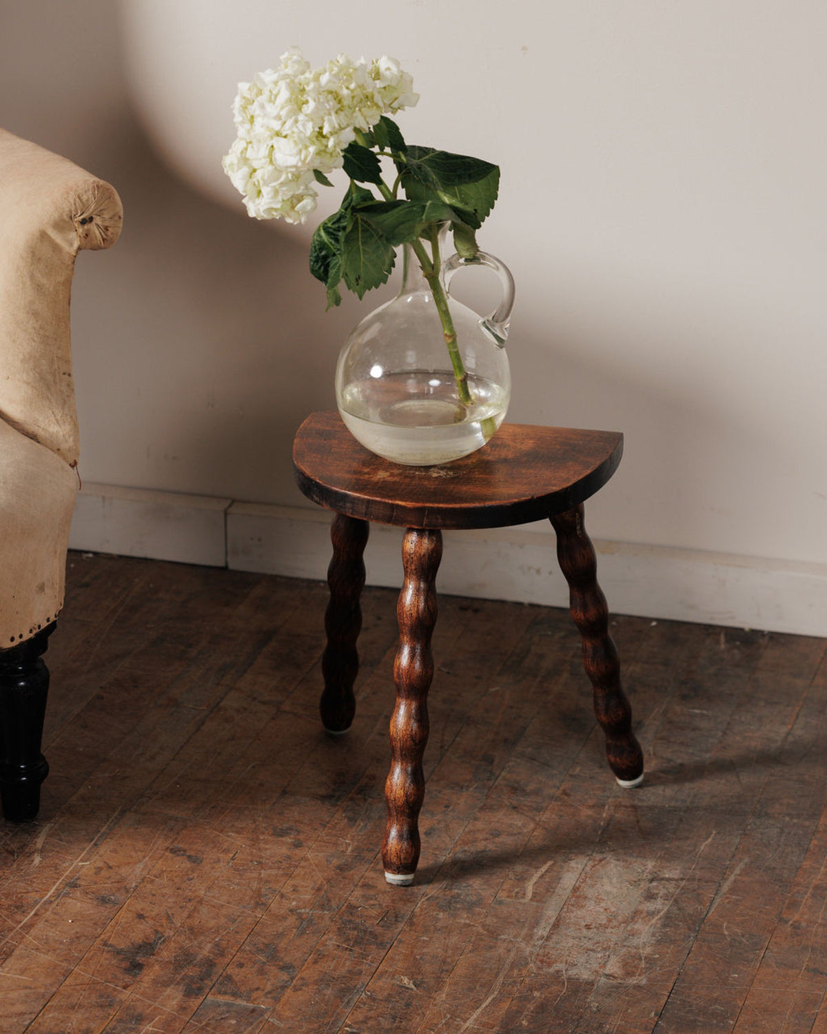 White hydrangeas in a clear jug vase on a dark wooden bobbin-leg stool.