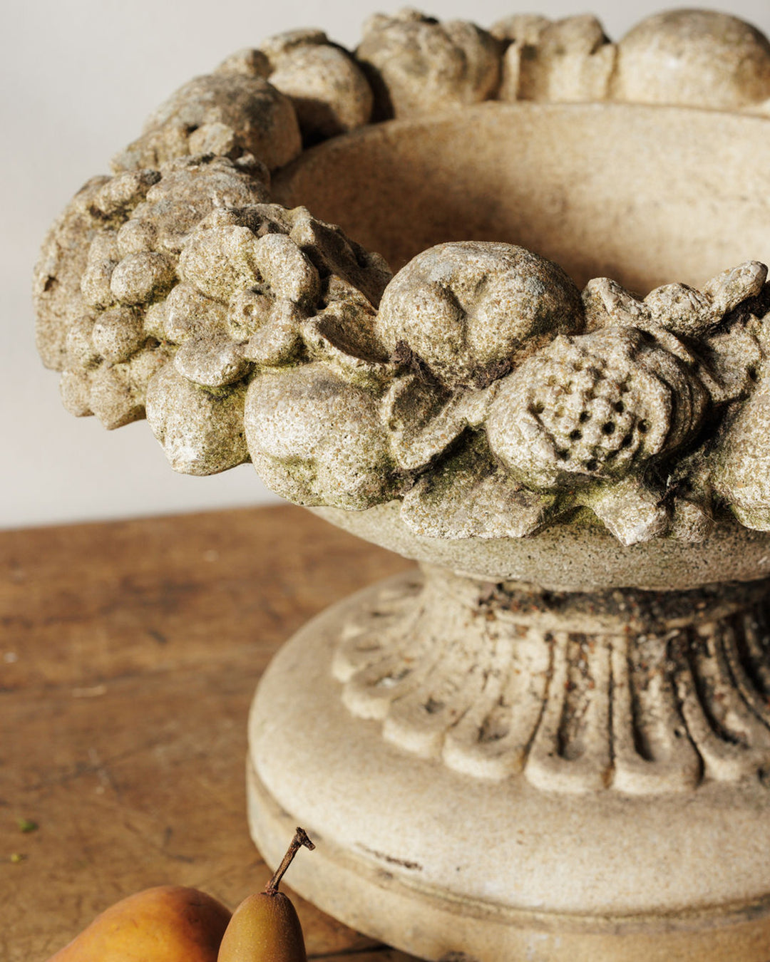 A rustic stone urn carved with fruit and flowers, on a wooden table with two pears.
