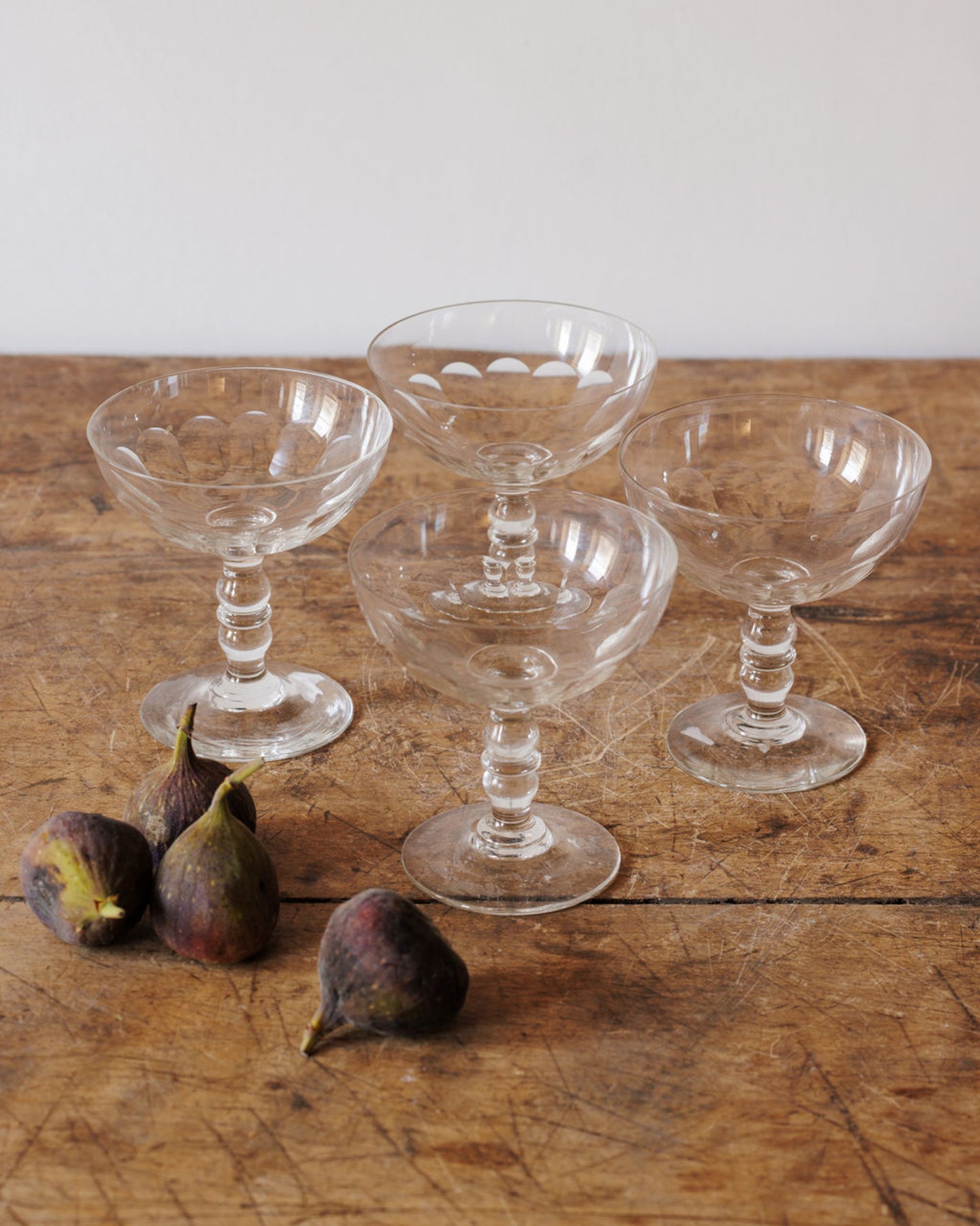 Four vintage glass coupes, one inverted, with a few fresh figs on a wooden table.