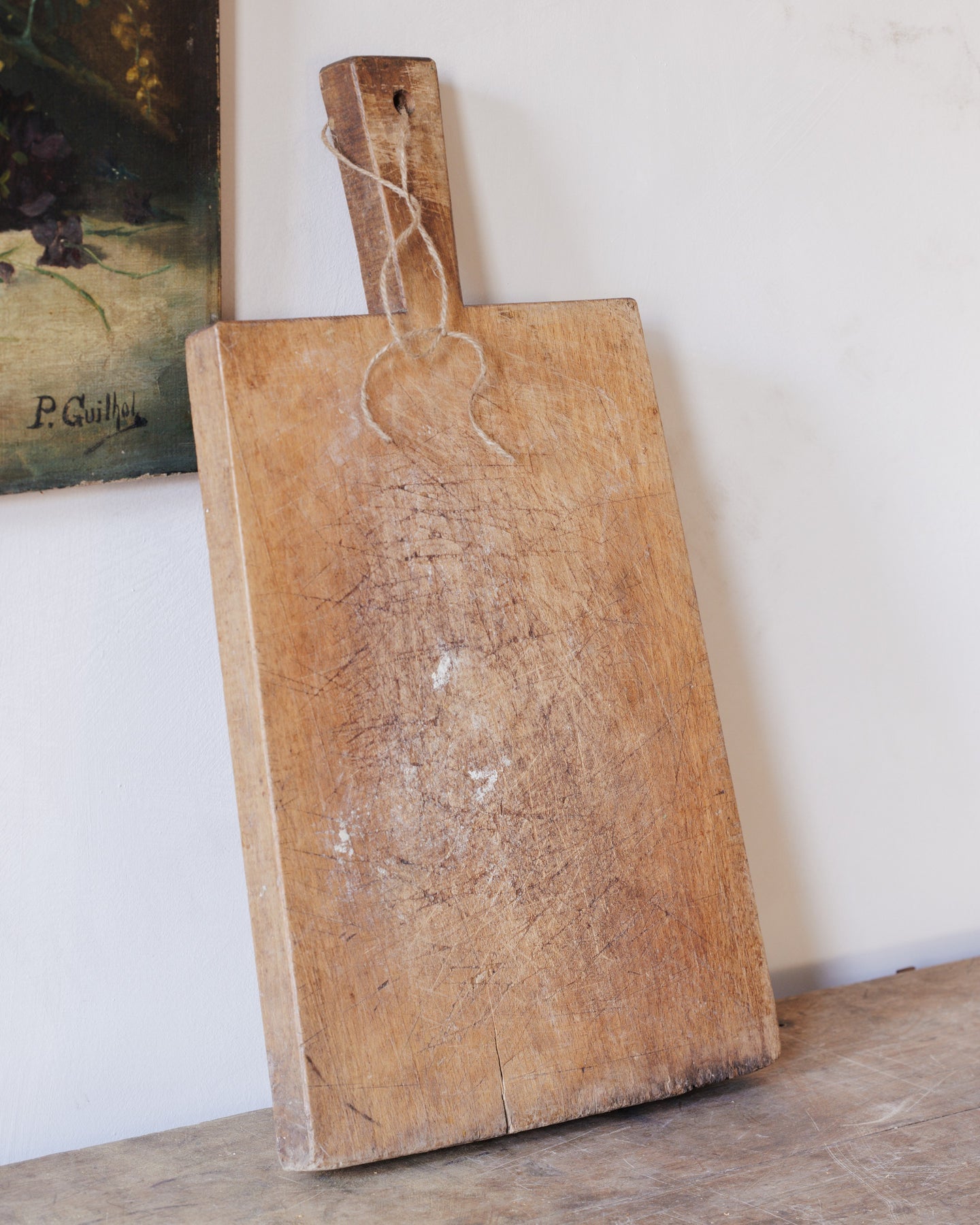 A well-worn wooden cutting board with a string loop on its handle leans against a white wall.